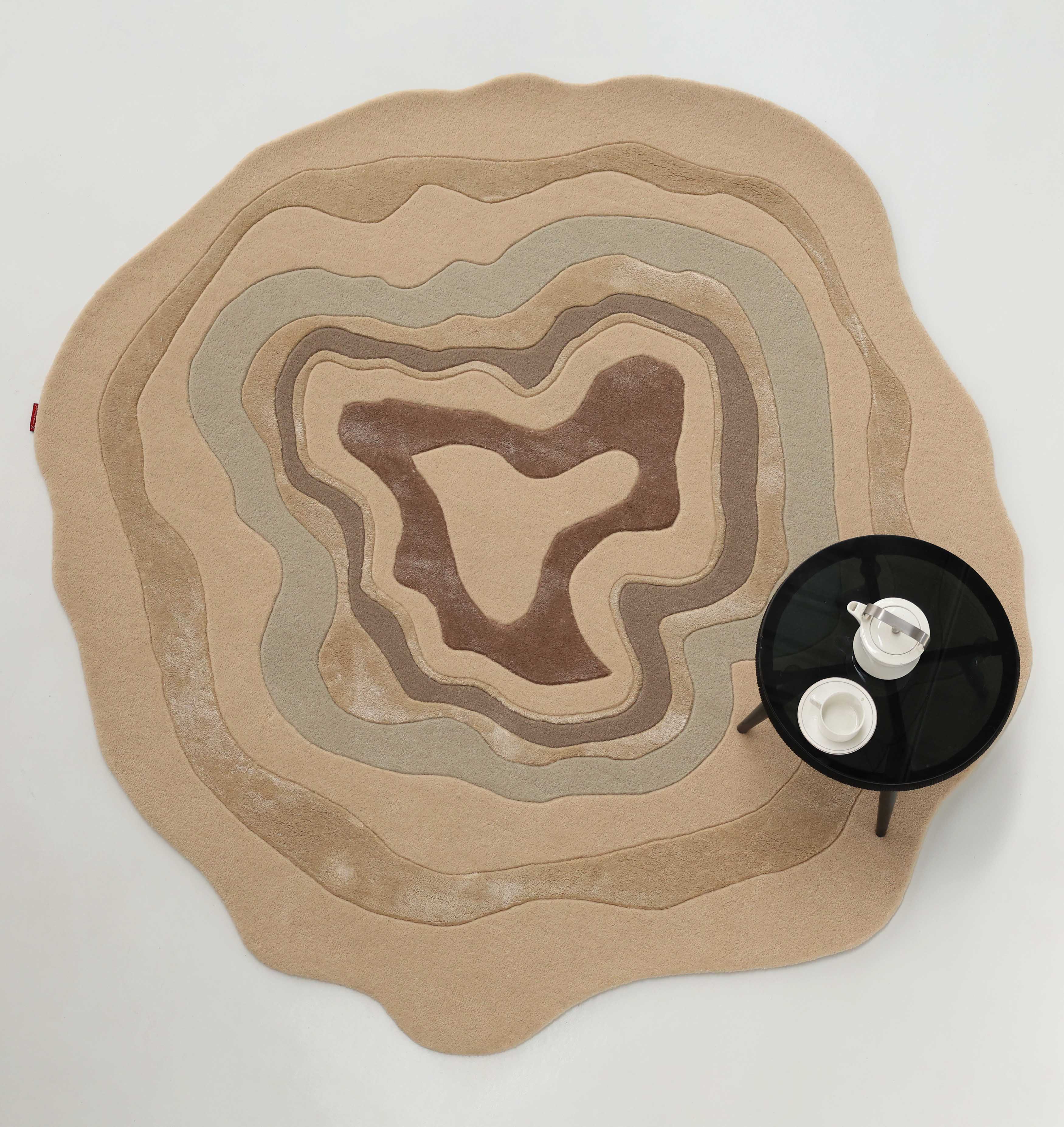 The RAMSHA HOME Luxury Abstract Contour Rug – Soft Textured Beige Accent Rug HTWV-194, with wavy organic shapes in beige and brown, is paired with a round black side table holding a teapot and cups on a white background.