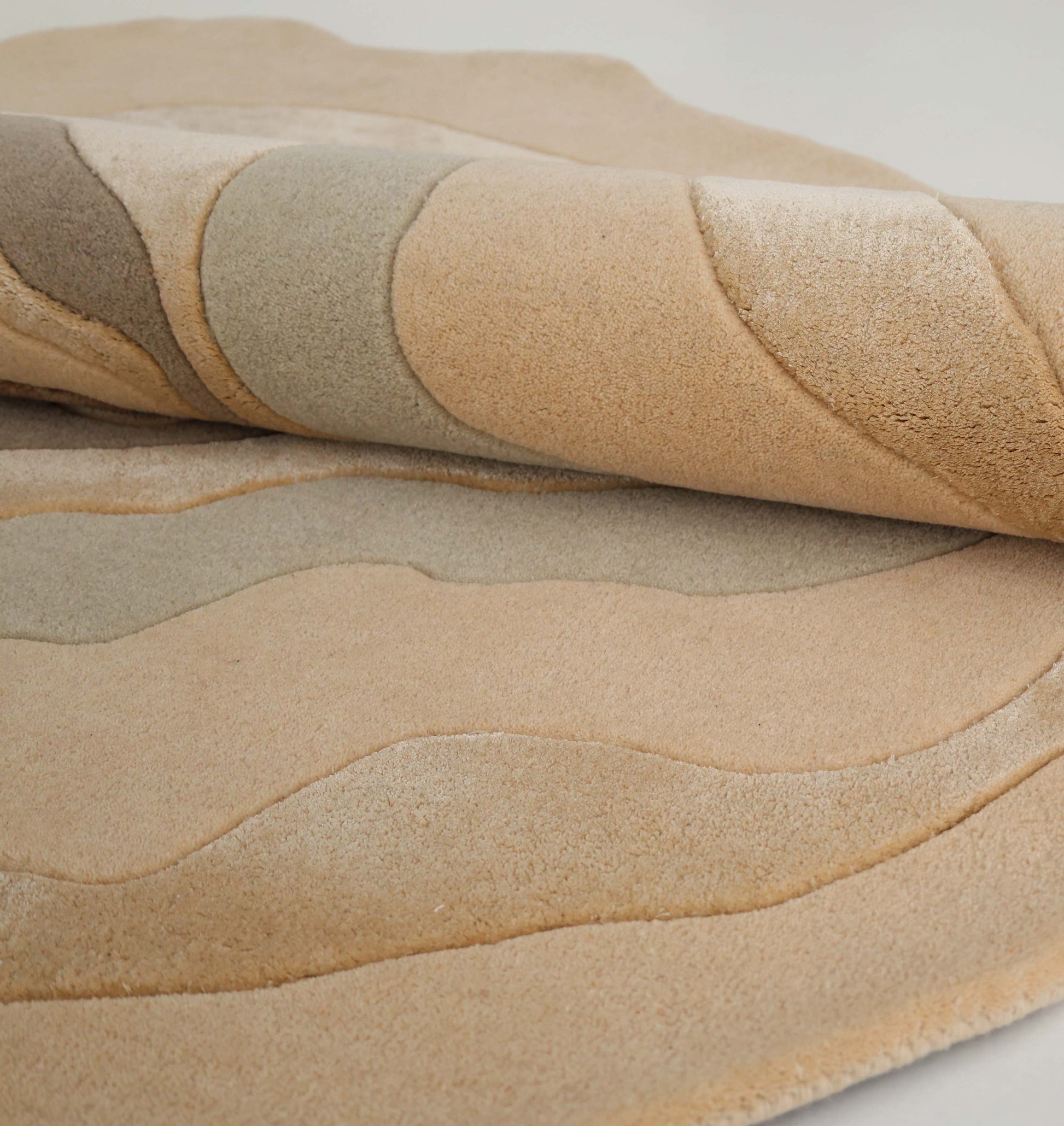 A close-up of the RAMSHA HOME Luxury Abstract Contour Rug HTWV-194, a soft, textured beige accent rug with wavy patterns, shown partially rolled on a white surface.