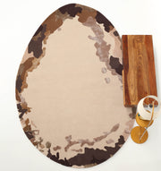 The RAMSHA HOME Terra Eclipse Oval Handmade Rug Accent Rug HTWV-195, featuring a camouflage-like brown, beige, and tan pattern, is placed beside a rectangular wooden table and a modern floor lamp on a white background.