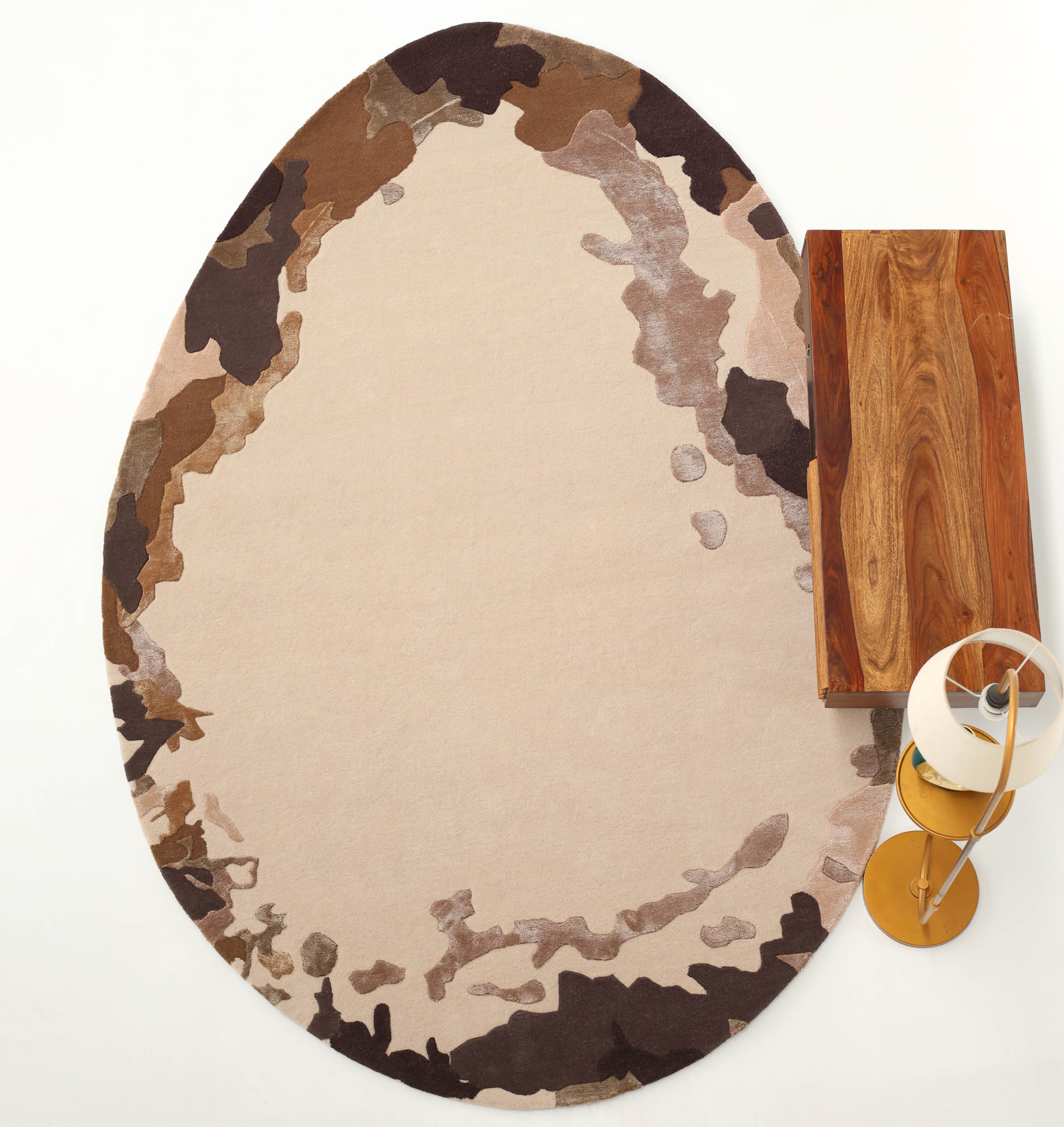 The RAMSHA HOME Terra Eclipse Oval Handmade Rug Accent Rug HTWV-195, featuring a camouflage-like brown, beige, and tan pattern, is placed beside a rectangular wooden table and a modern floor lamp on a white background.