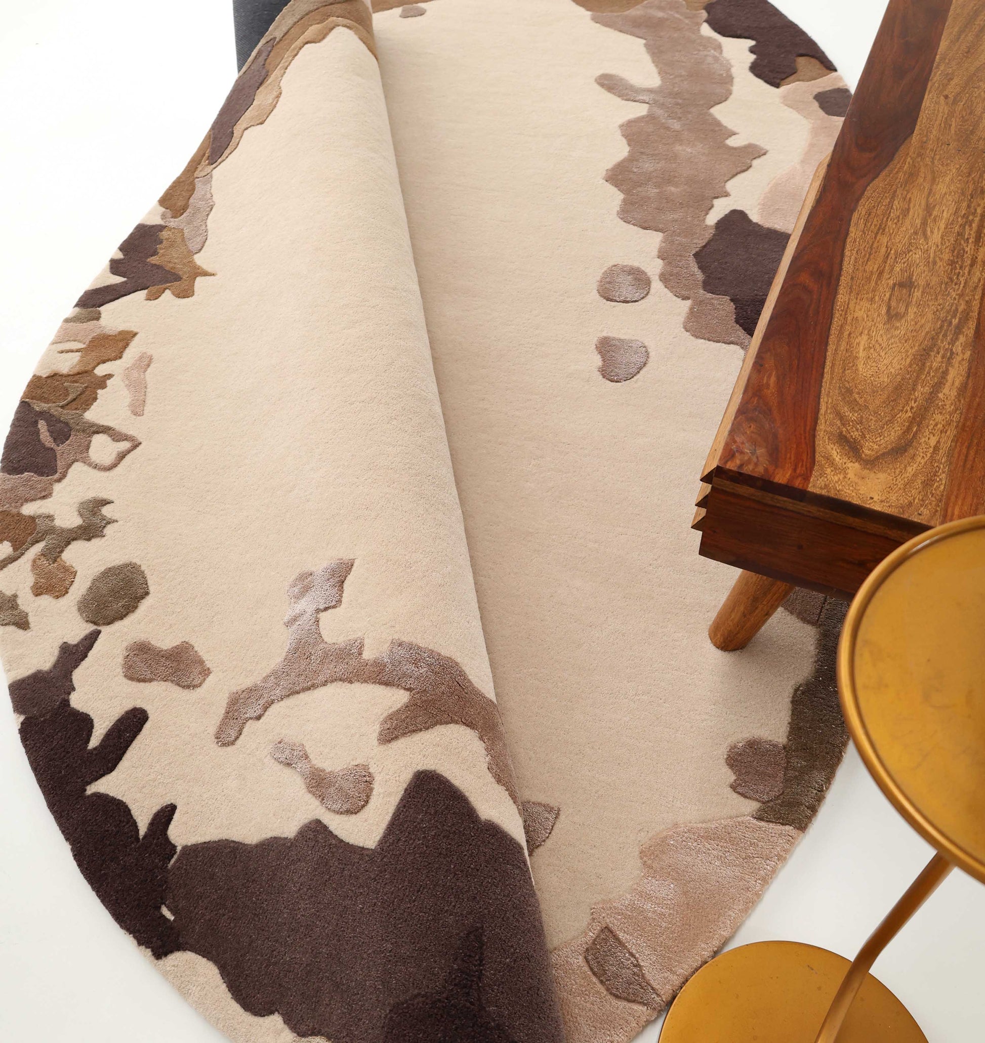 The RAMSHA HOME Terra Eclipse Oval Handmade Rug Accent Rug HTWV-195 with abstract brown, beige, and cream patterns is partially folded near a wooden table and gold metal chair on a white floor.