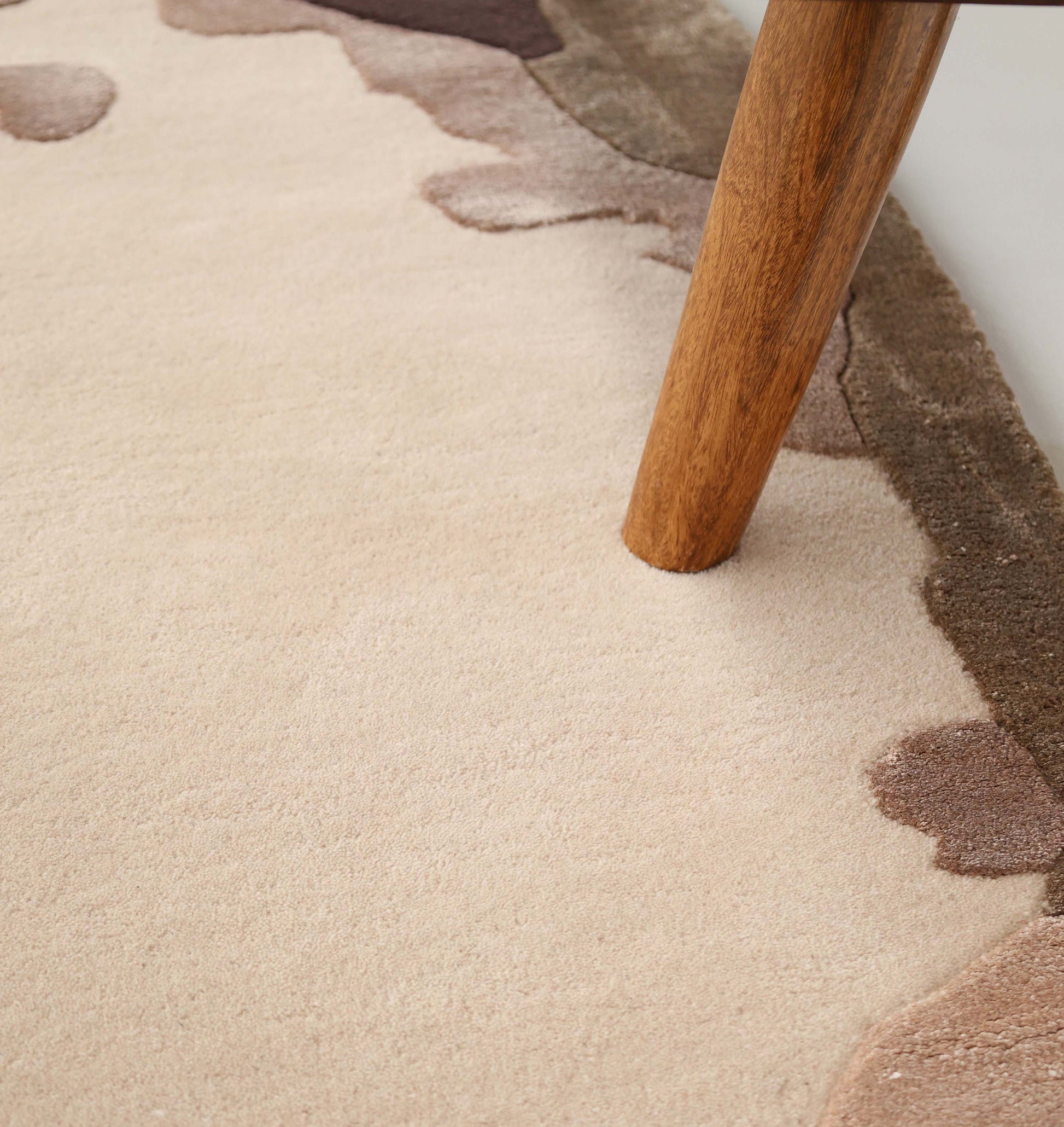 A close-up of the RAMSHA HOME Terra Eclipse Oval Handmade Rug Accent Rug HTWV-195 in beige and brown abstract shapes, with a wooden furniture leg resting on it.