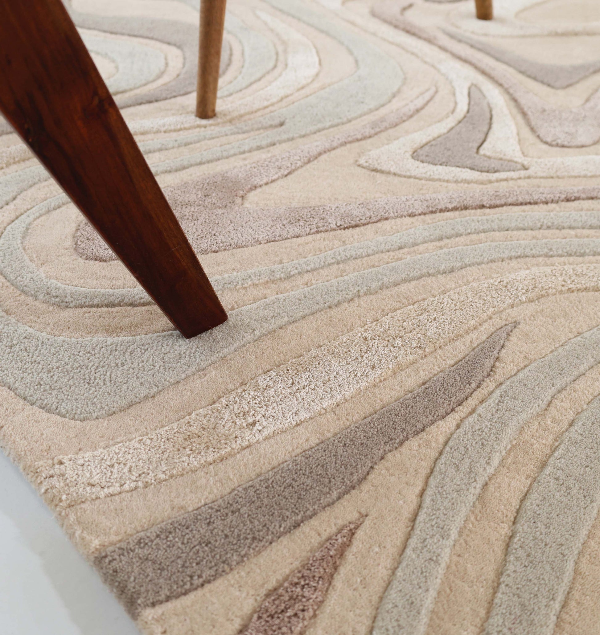 Close-up of the RAMSHA HOME Desginer Freeform Handmade Rug HTWV-196, featuring beige, cream, and light brown wavy abstract patterns. A dark wooden furniture leg rests partly on this modern interior rug.