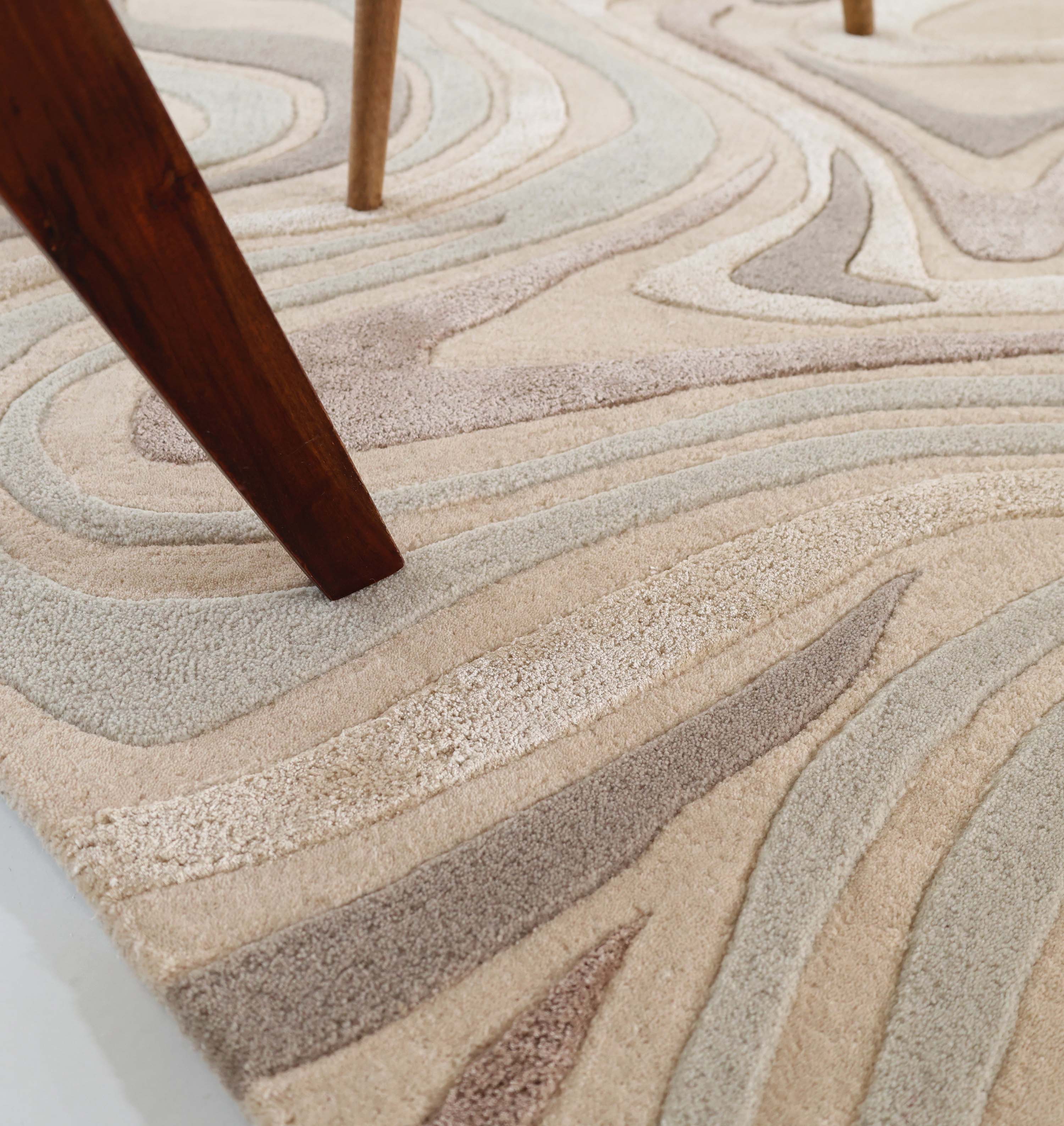 Close-up of the RAMSHA HOME Desginer Freeform Handmade Rug HTWV-196, featuring beige, cream, and light brown wavy abstract patterns. A dark wooden furniture leg rests partly on this modern interior rug.