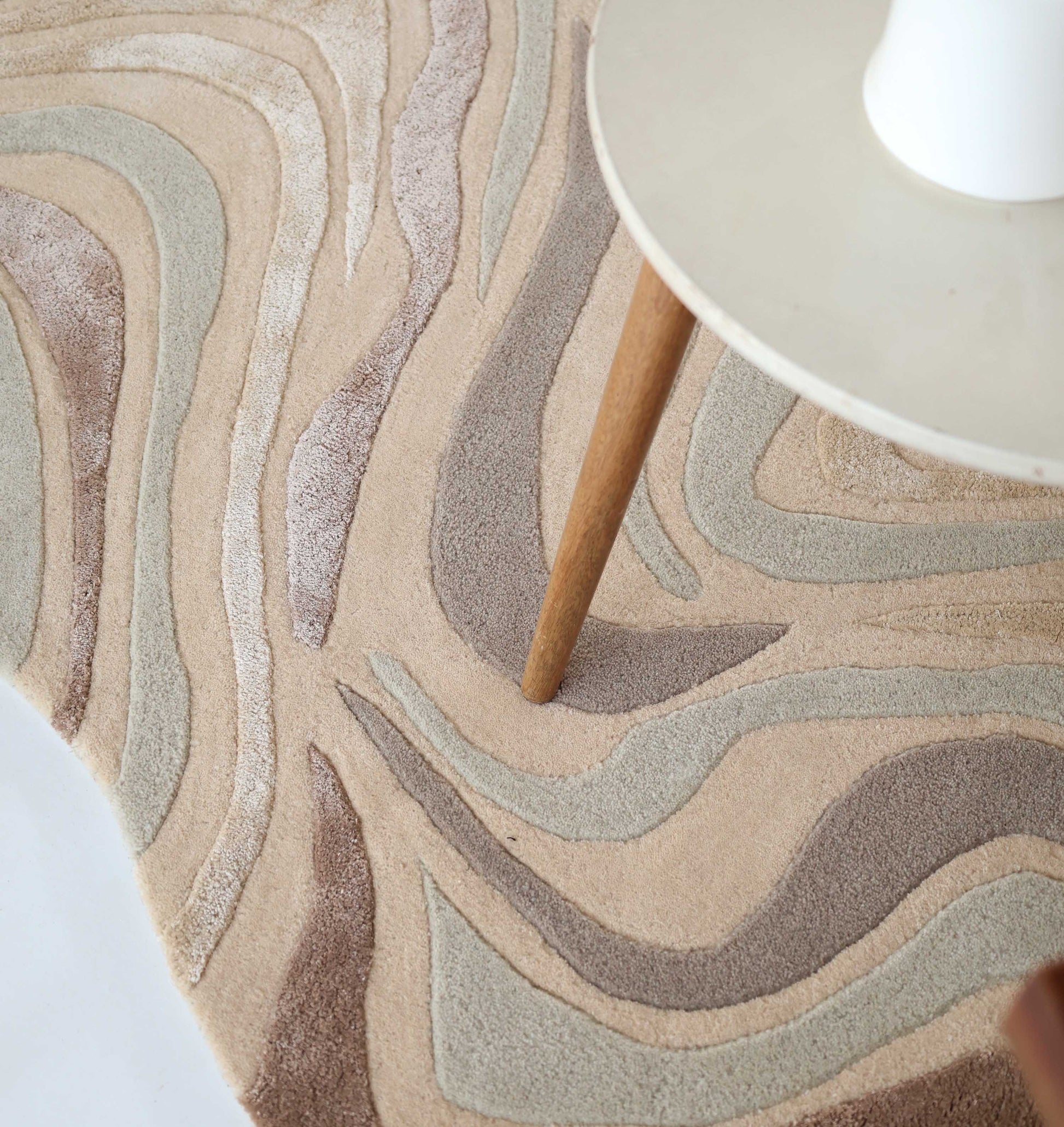 A close-up of the RAMSHA HOME Designer Freeform Handmade Rug HTWV-196, a beige and pastel abstract rug with cream, light gray, and taupe wavy shapes, under a round table with wooden legs in a modern interior.