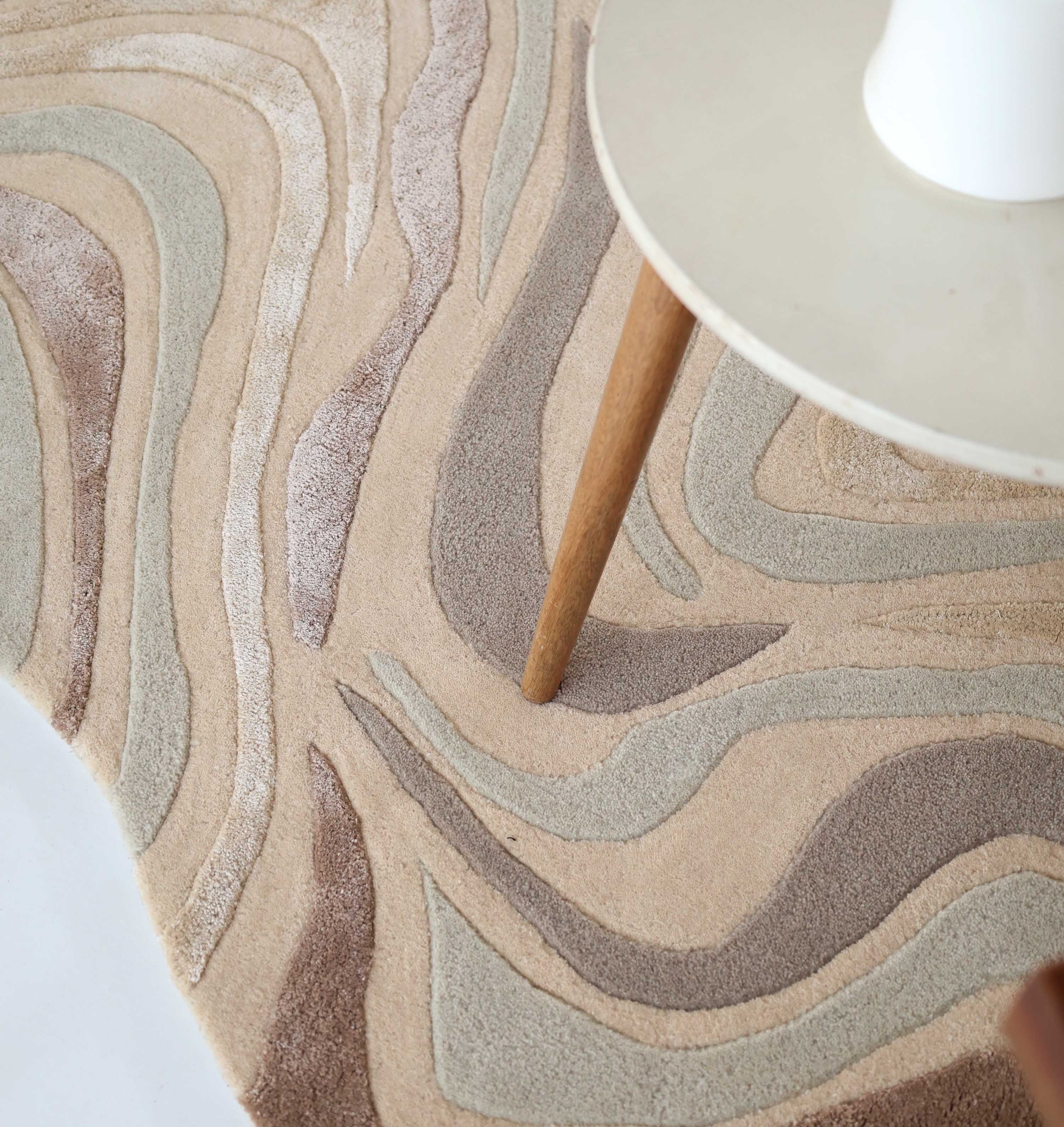 A close-up of the RAMSHA HOME Designer Freeform Handmade Rug HTWV-196, a beige and pastel abstract rug with cream, light gray, and taupe wavy shapes, under a round table with wooden legs in a modern interior.
