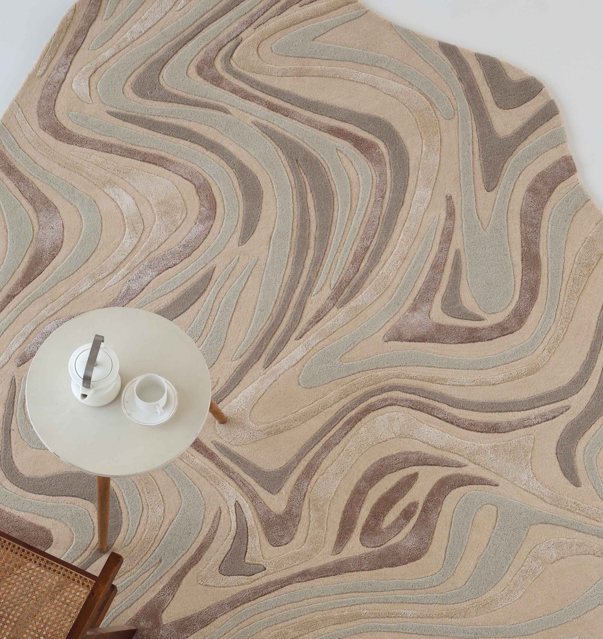 The RAMSHA HOME Desginer Freeform Handmade Rug HTWV-196, featuring a wavy abstract pattern in beige, brown, and gray, is placed beside a round table with tea settings and part of a woven chair visible.