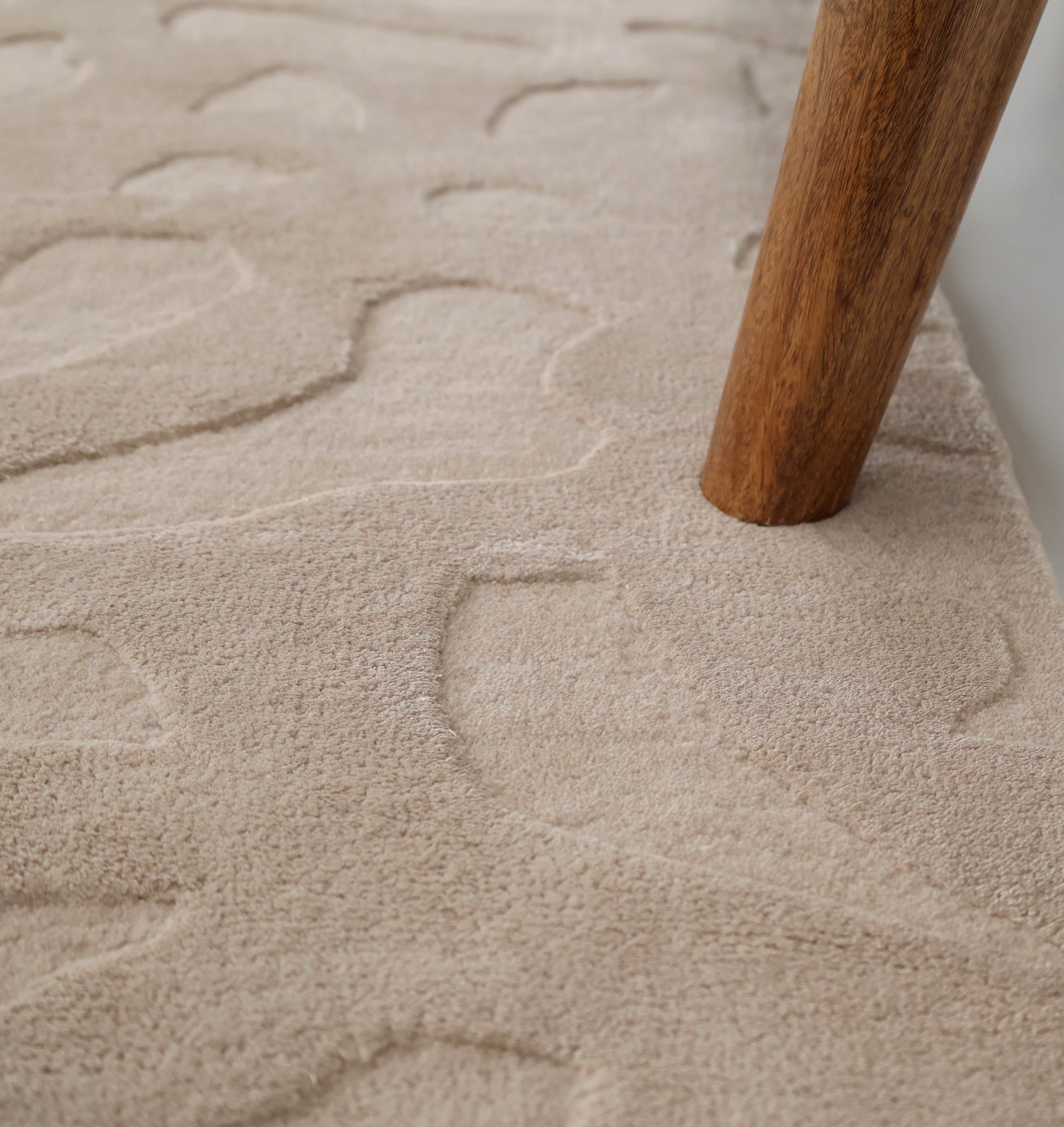 Close-up of a RAMSHA HOME Rectangle Modern Handmade Rug 200x300 CM HTWV-197, featuring a beige textured surface with embossed abstract bird shapes, displayed next to a wooden furniture leg.
