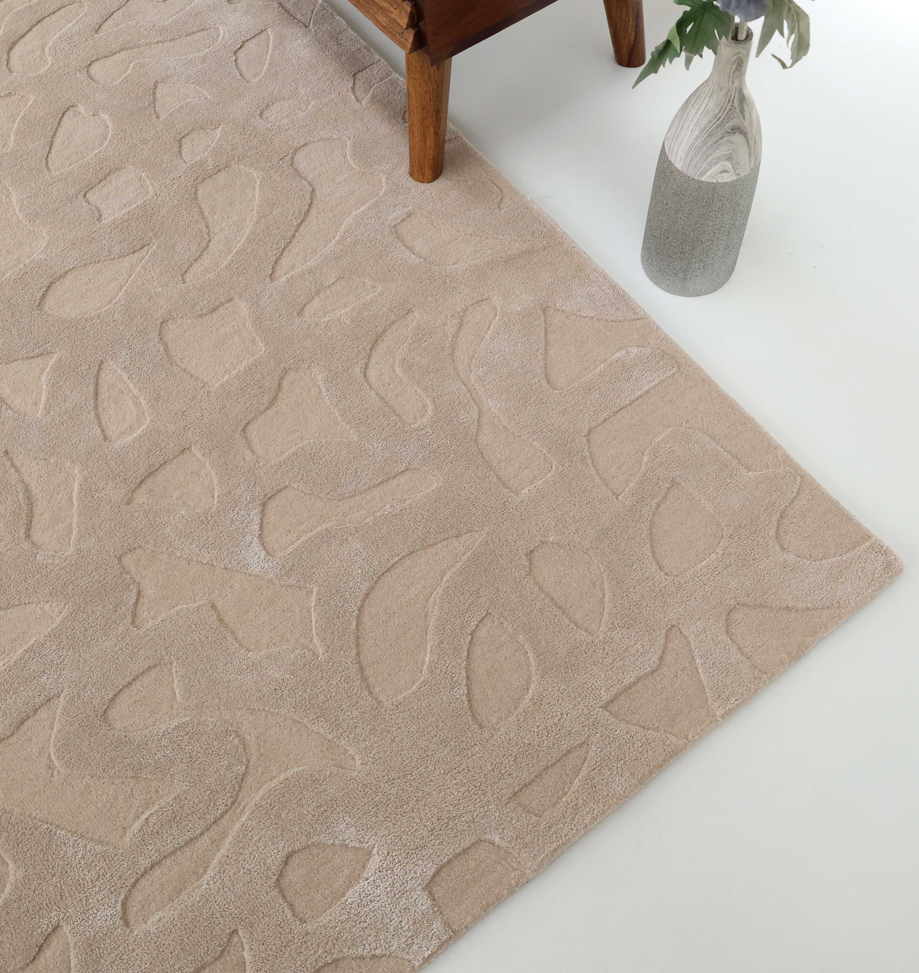 The RAMSHA HOME Rectangle Modern Handmade Rug 200x300 CM HTWV-197 features beige, abstract patterns and a raised texture, shown beside a wooden furniture leg and a vase with green leaves on a white floor.