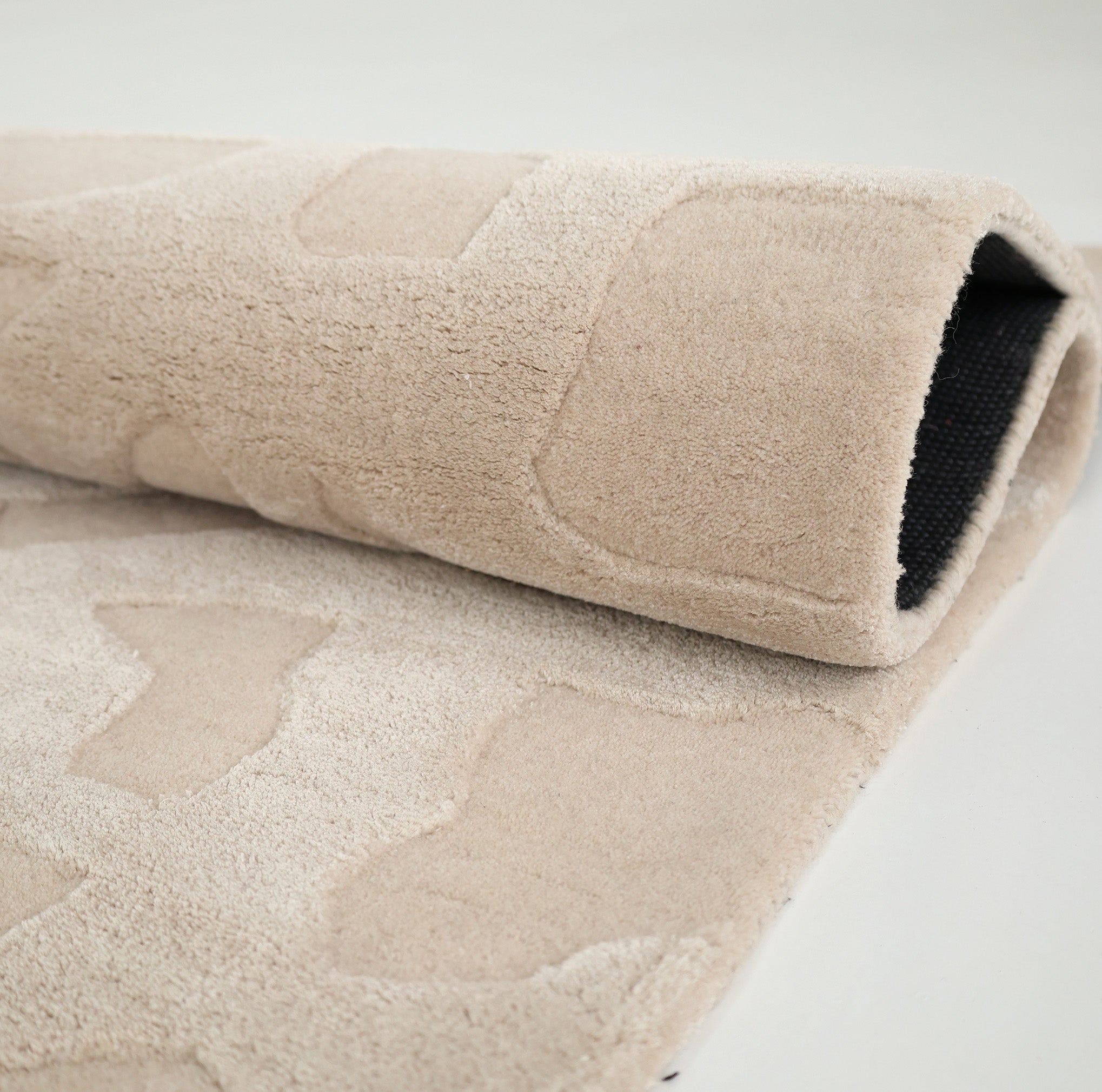 A close-up of the RAMSHA HOME Rectangle Modern Handmade Rug 200x300 CM HTWV-197, beige with abstract raised patterns, partially rolled up to show its black non-slip backing on a light surface.