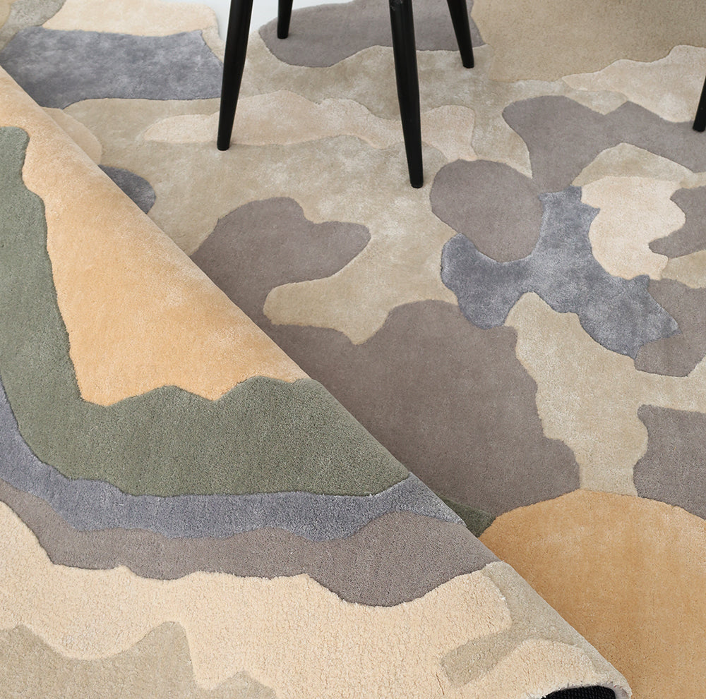 The RAMSHA HOME Contemporary Flow Area Handmade Rug HTWV-199 features an abstract camouflage design in beige, tan, green, and gray, shown partially rolled under black chair legs—a striking modern interior accent.