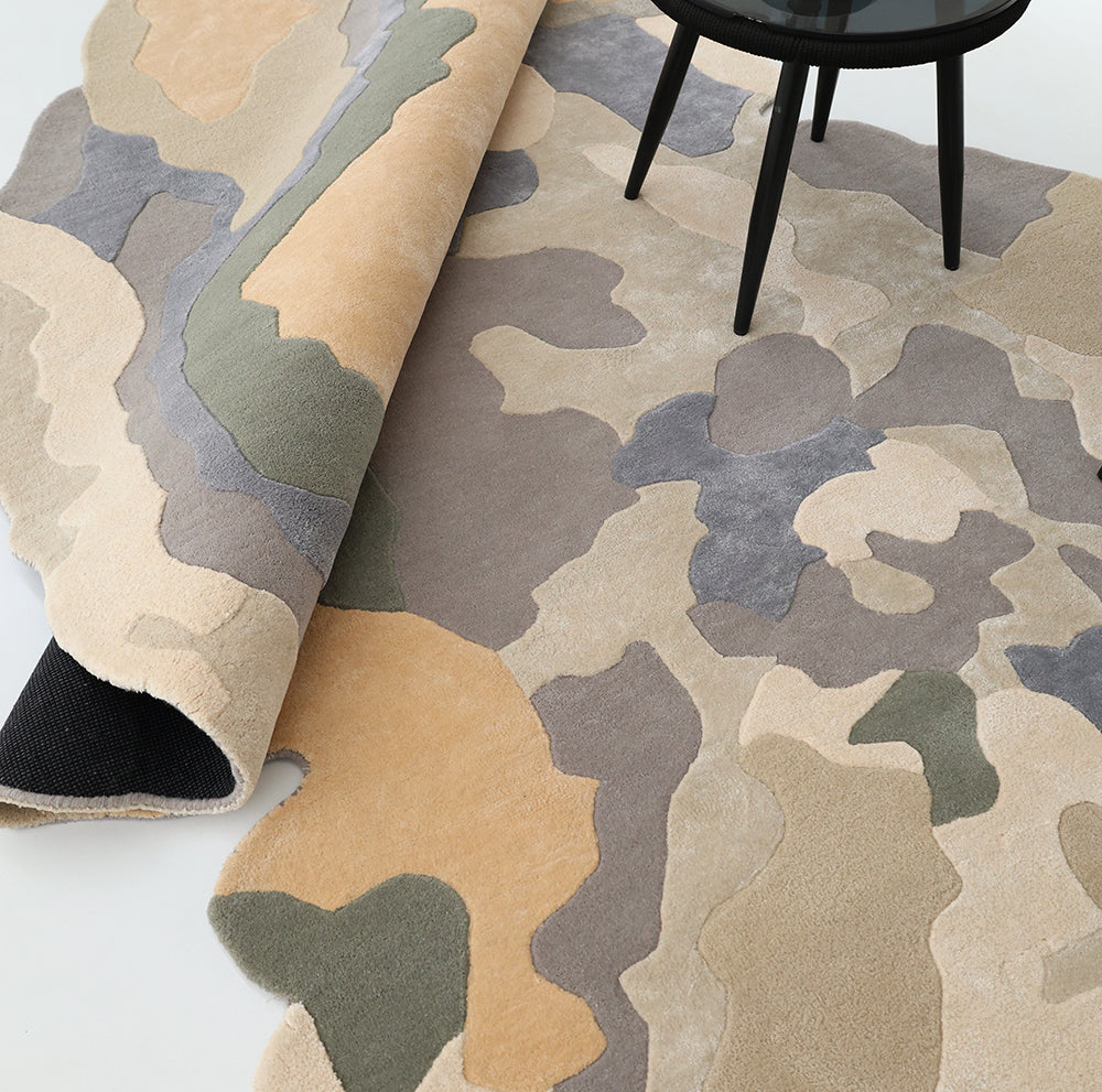 A close-up of the RAMSHA HOME Contemporary Flow Area Handmade Rug HTWV-199, featuring an abstract camouflage pattern in beige, tan, olive, gray, and cream tones, partially rolled with a black-legged table on top.