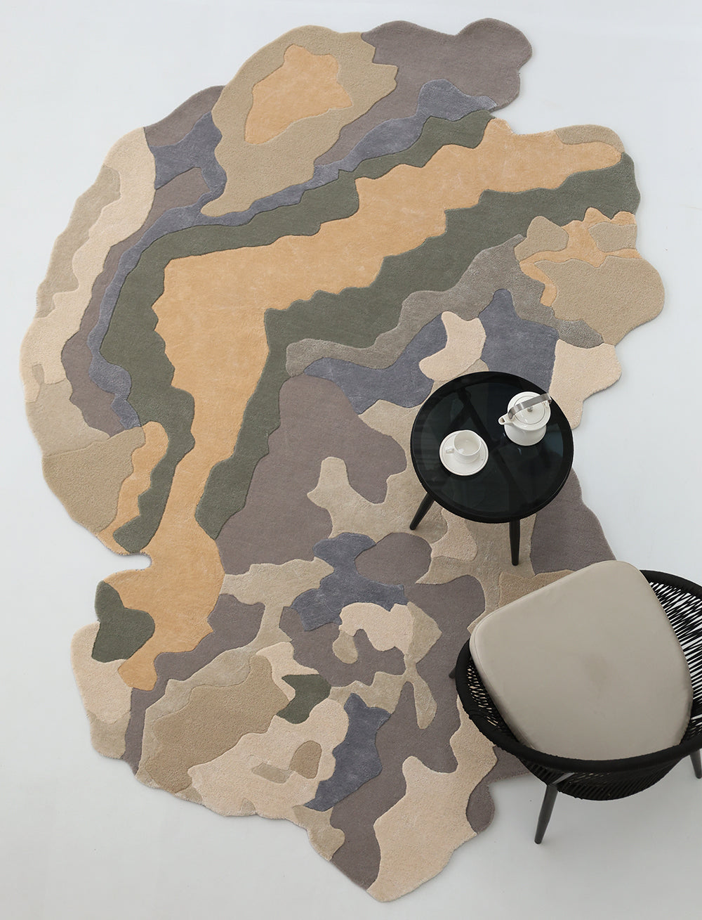 The Contemporary Flow Area Handmade Rug HTWV-199 by RAMSHA HOME, featuring abstract wavy shapes in beige, tan, olive, gray, and sage, is displayed on a white floor beside a round black table and a woven chair with a light cushion.