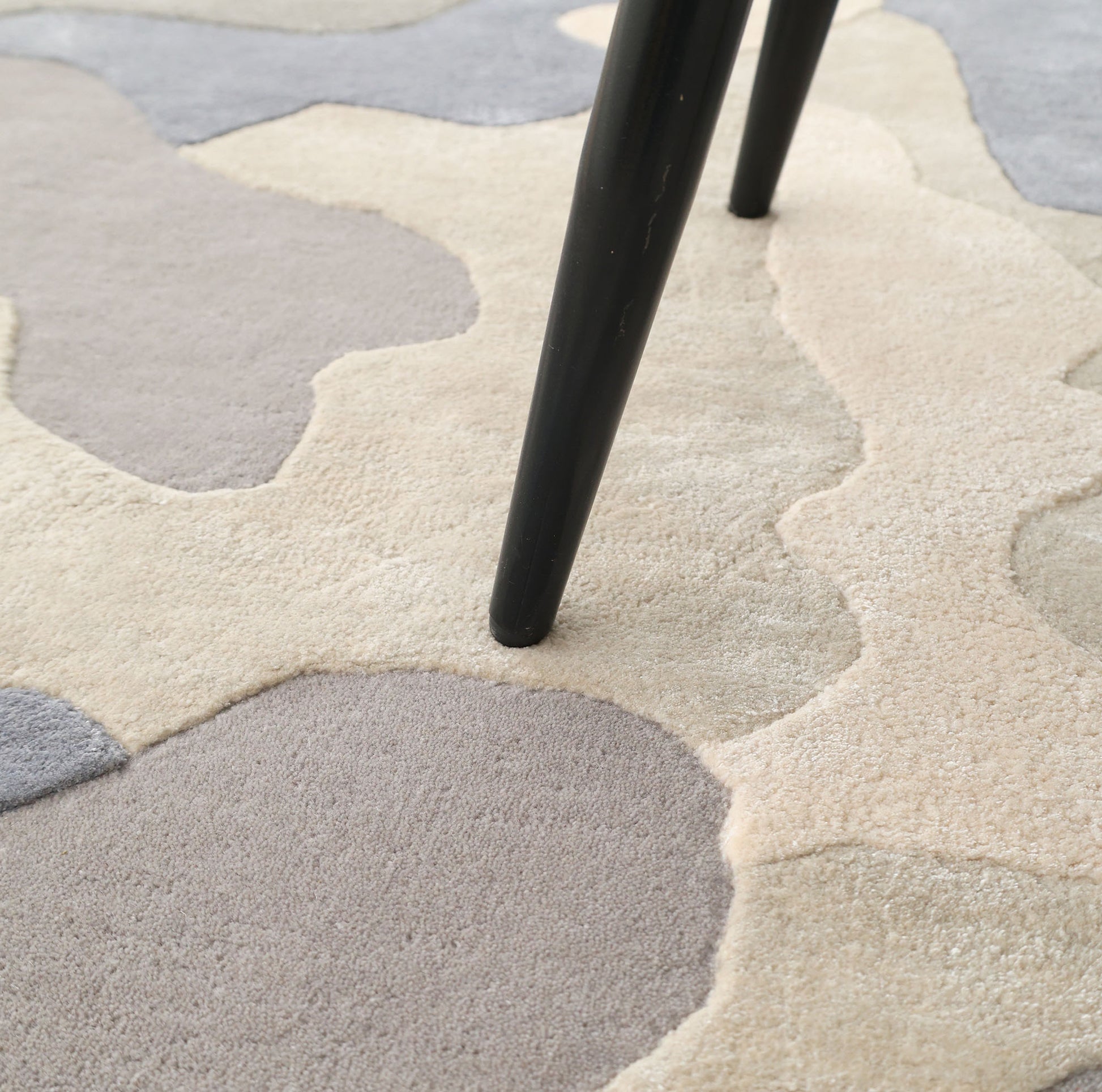 A close-up of a black table or chair leg on the RAMSHA HOME Contemporary Flow Area Handmade Rug HTWV-199, featuring irregular abstract patches in beige, gray, and cream.