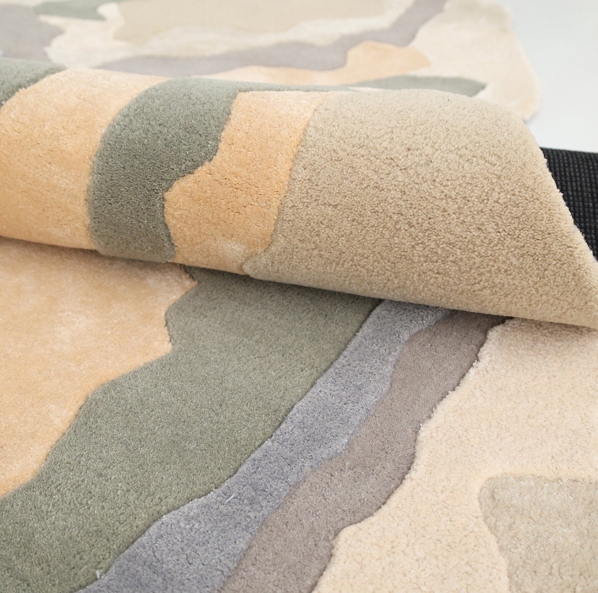 A close-up of the RAMSHA HOME Contemporary Flow Area Handmade Rug HTWV-199 shows its wavy beige, green, yellow, and gray design, with a rolled corner revealing textured crafting, a black underside, and its unique abstract border style.