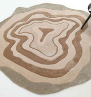 The RAMSHA HOME Desginer Freeform Handmade Rug HTWV-200 features layered, concentric beige and brown patterns resembling topographic lines; a black chair is partially visible at the rug's abstract edge.