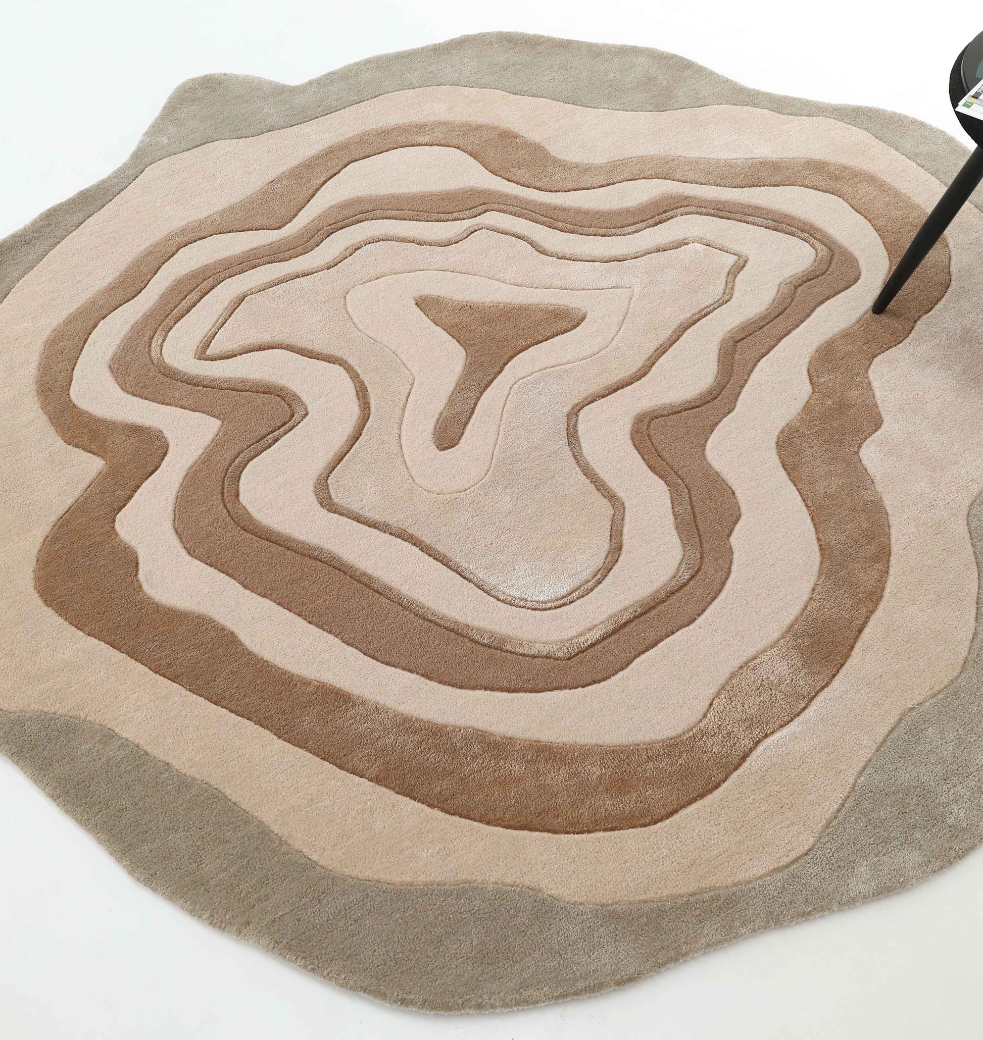 The RAMSHA HOME Desginer Freeform Handmade Rug HTWV-200 features layered, concentric beige and brown patterns resembling topographic lines; a black chair is partially visible at the rug's abstract edge.