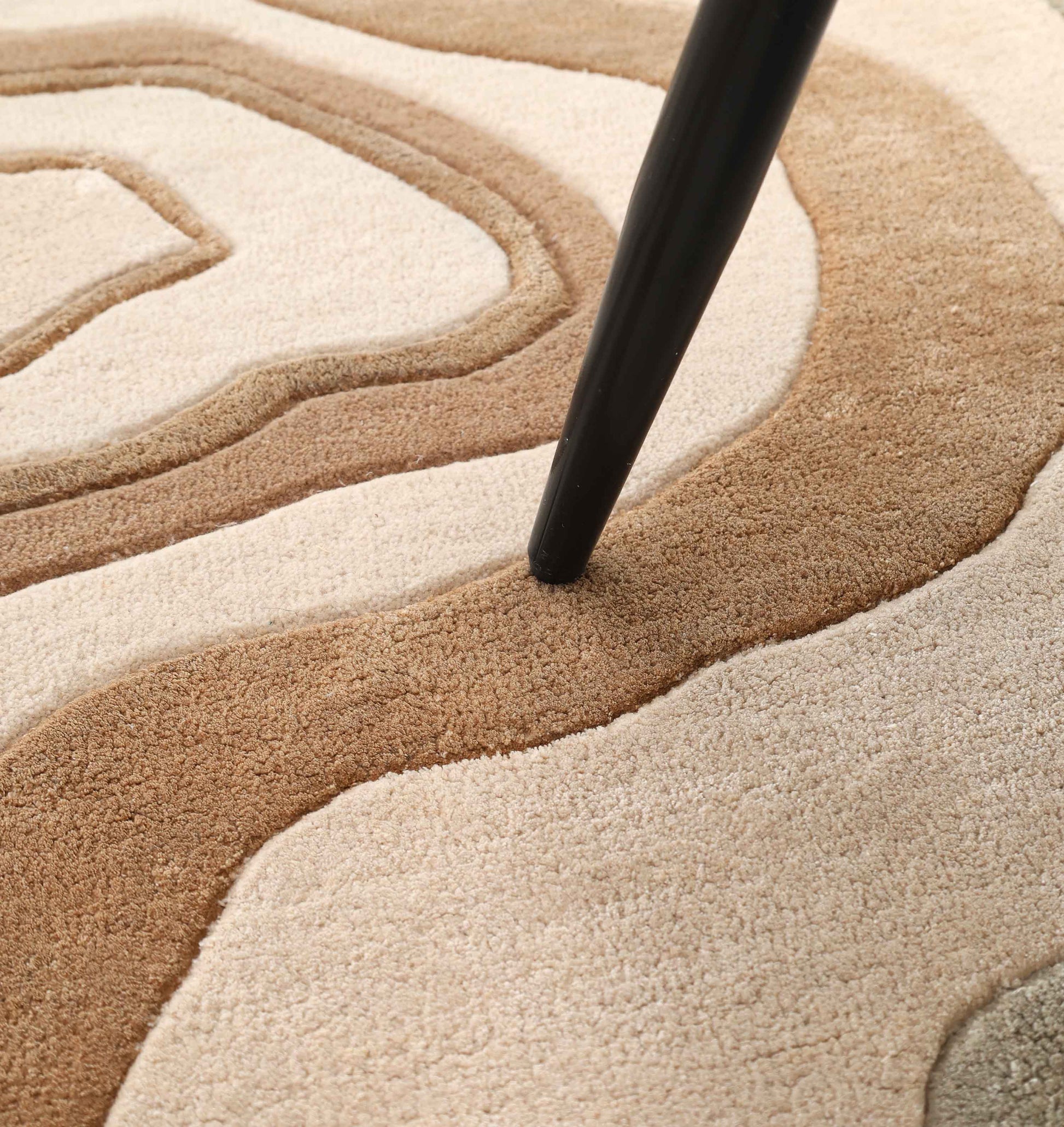 A black tapered furniture leg rests on the RAMSHA HOME Designer Freeform Handmade Rug HTWV-200, featuring a textured, wavy pattern in beige, tan, and brown.