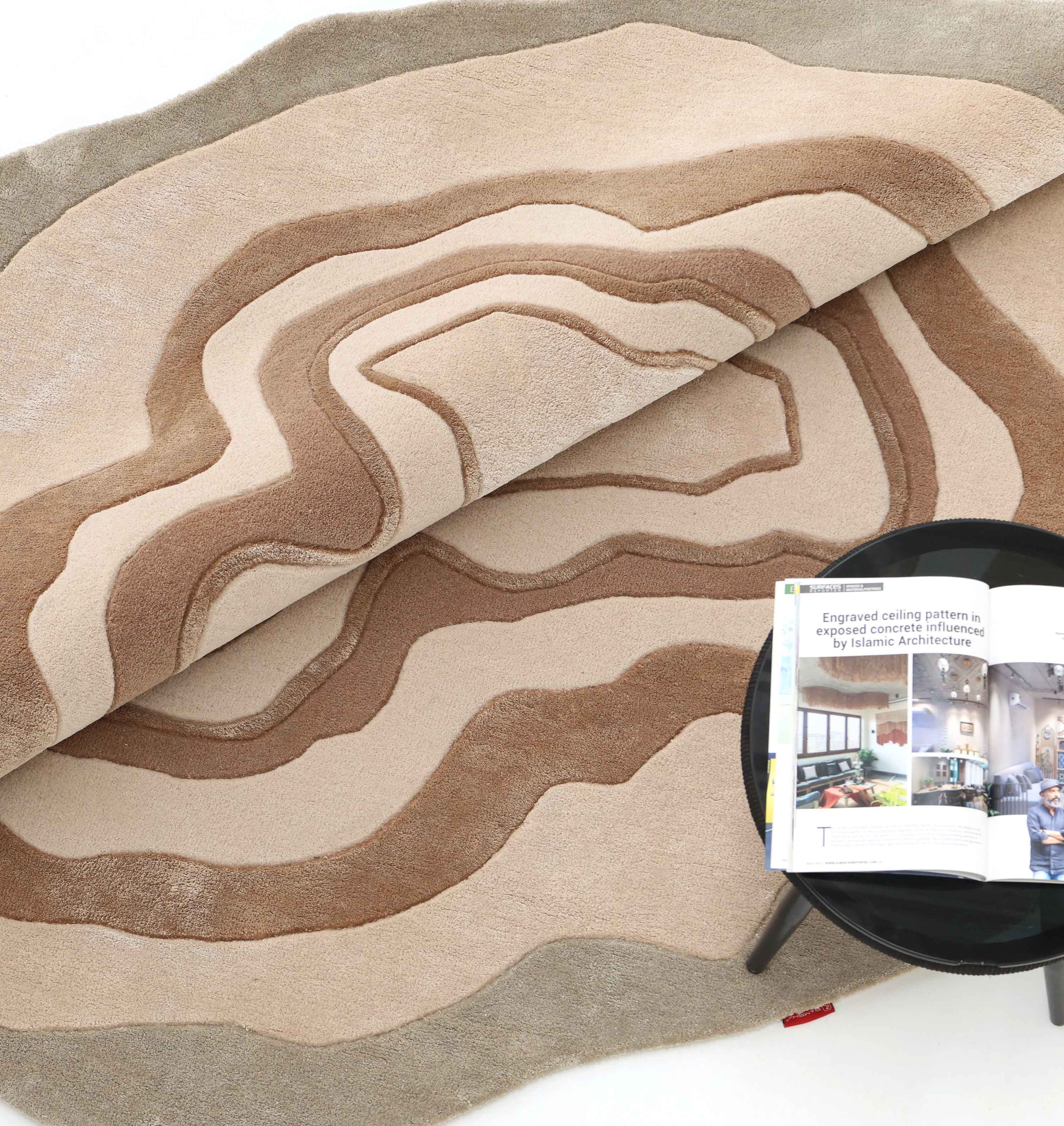 The RAMSHA HOME Desginer Freeform Handmade Rug HTWV-200, featuring earth-tone topographic patterns, is partially rolled back beside a round black table topped with an open magazine displaying interior design photos.