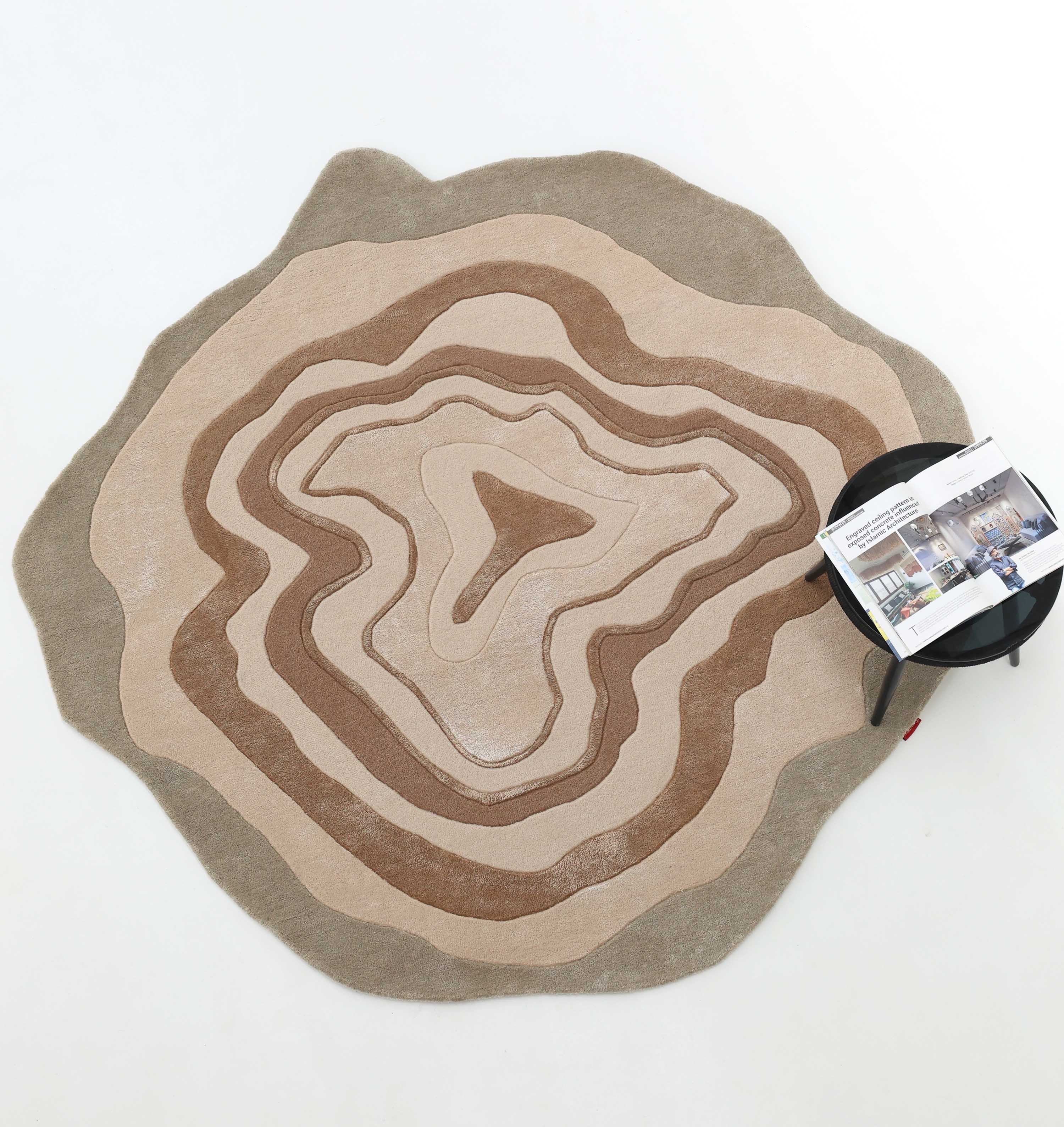 The RAMSHA HOME Desginer Freeform Handmade Rug HTWV-200 features earth-toned, wavy patterns like tree rings and sits on a white floor beside a black side table with magazines—a striking modern accent.