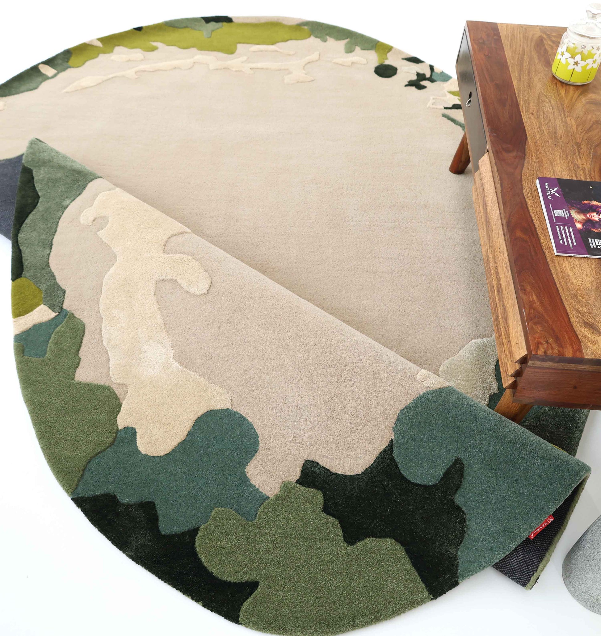 The RAMSHA HOME Terra Eclipse Oval Handmade Rug HTWV-201, featuring an abstract beige and green pattern, is partially folded near a wooden coffee table with a yellow candle and magazines.