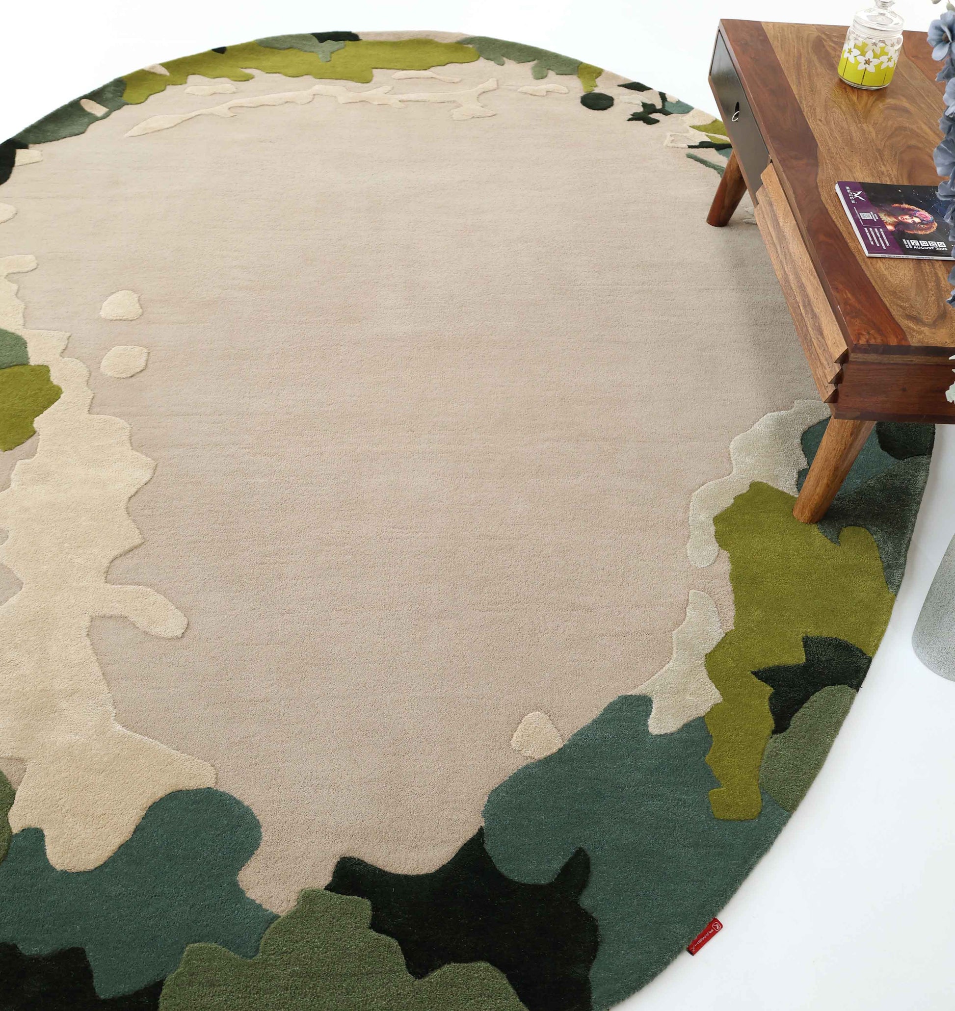 The RAMSHA HOME Terra Eclipse Oval Handmade Rug HTWV-201, featuring an abstract design in beige, green, and olive tones, is placed under a wooden table holding magazines, a candle, and a small plant.
