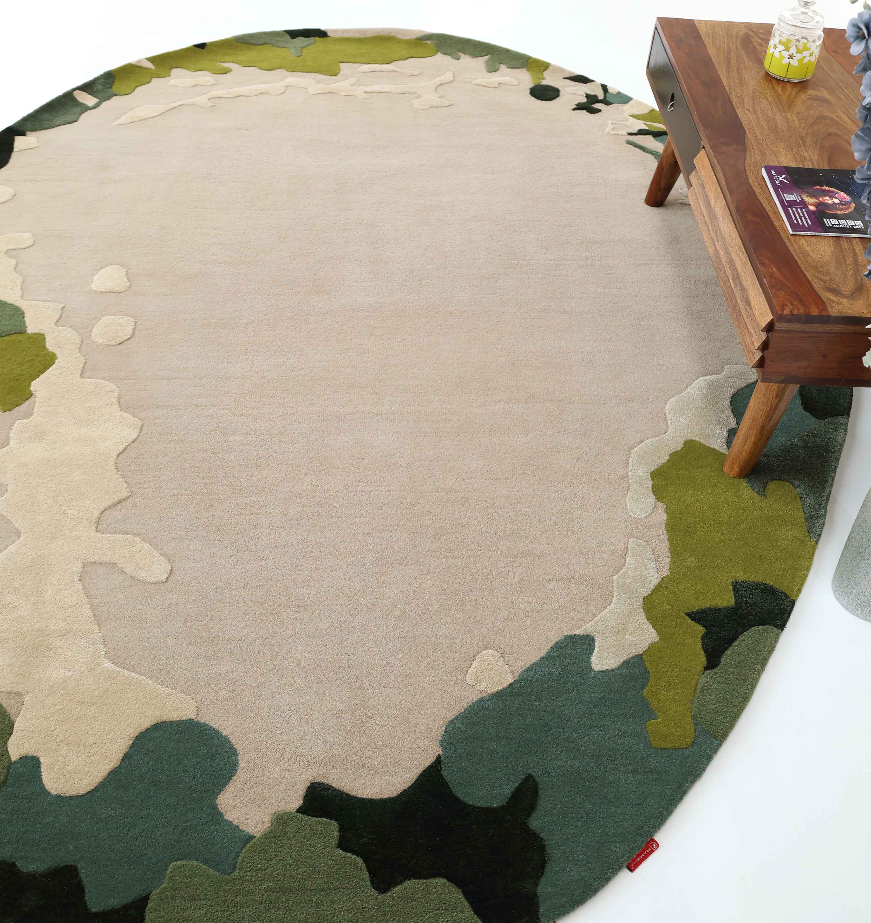 The RAMSHA HOME Terra Eclipse Oval Handmade Rug HTWV-201, featuring an abstract design in beige, green, and olive tones, is placed under a wooden table holding magazines, a candle, and a small plant.