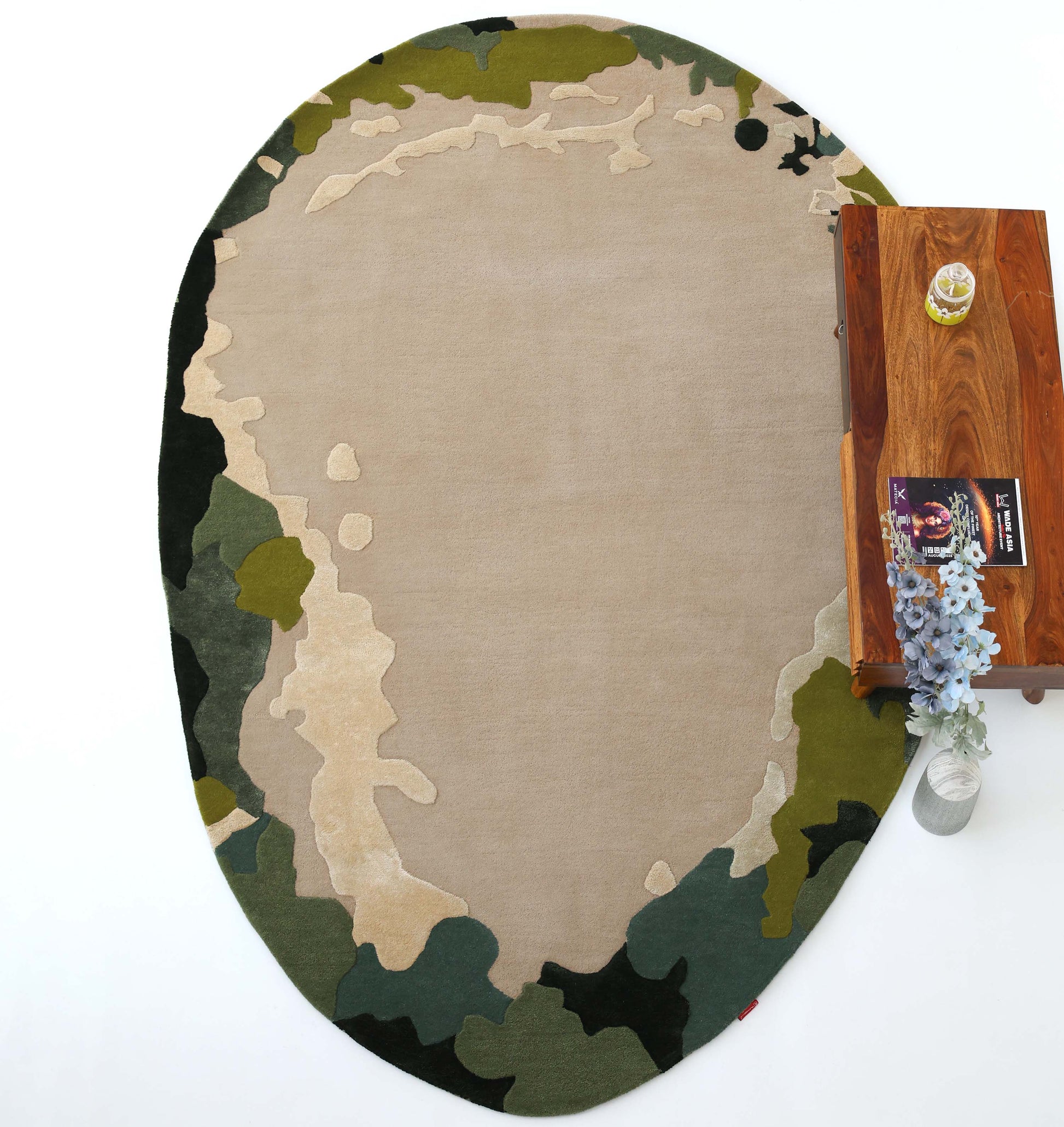 The Terra Eclipse Oval Handmade Rug HTWV-201 by RAMSHA HOME, featuring a green, beige, and brown camouflage pattern, lies on a white floor next to a wooden table—an ideal modern accent for contemporary interiors.