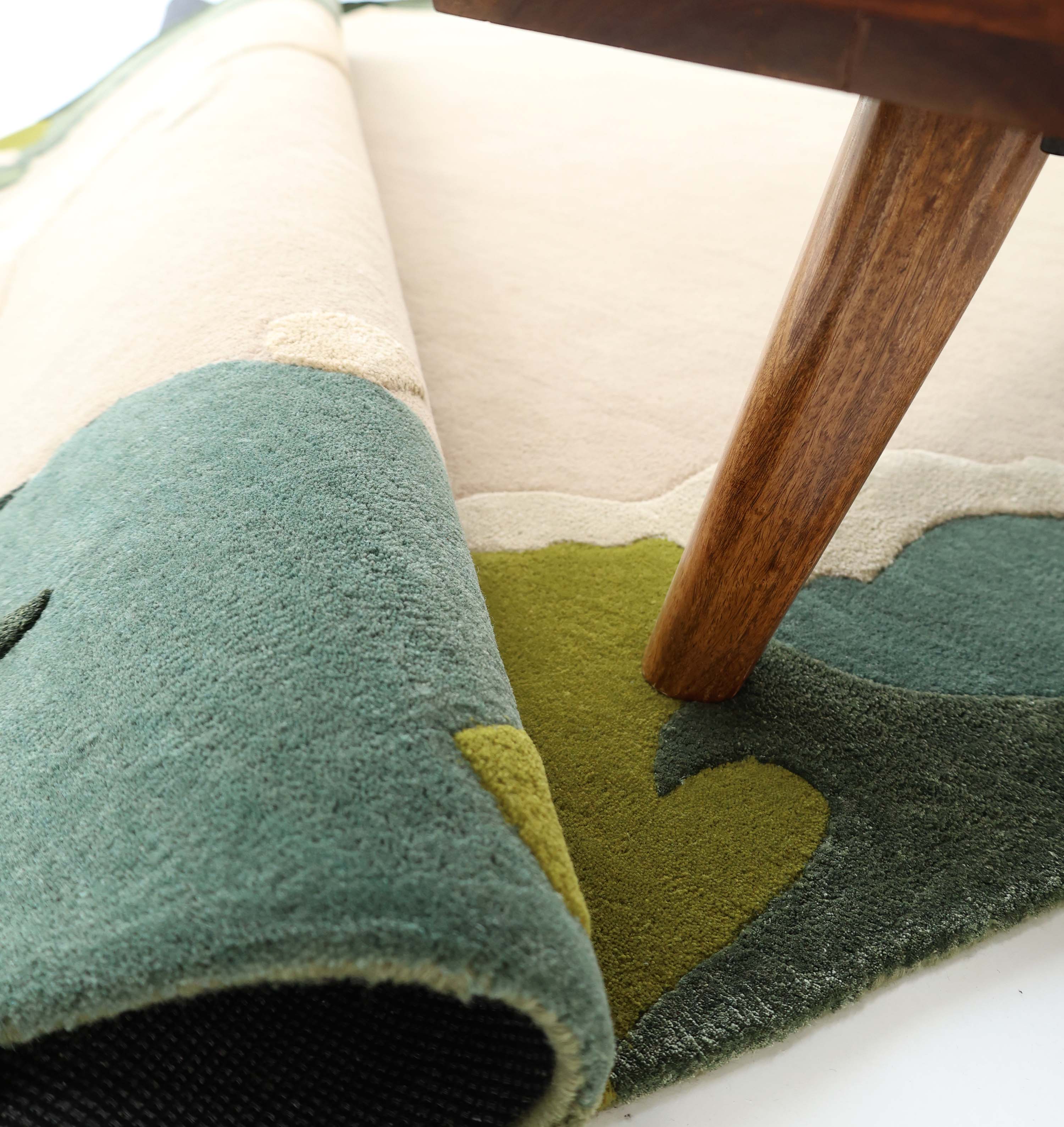 A close-up of the RAMSHA HOME Terra Eclipse Oval Handmade Rug Accent Rug HTWV-201, featuring green, olive, and cream abstract patterns. A wooden table leg rests on the rug, highlighting its thickness and texture.