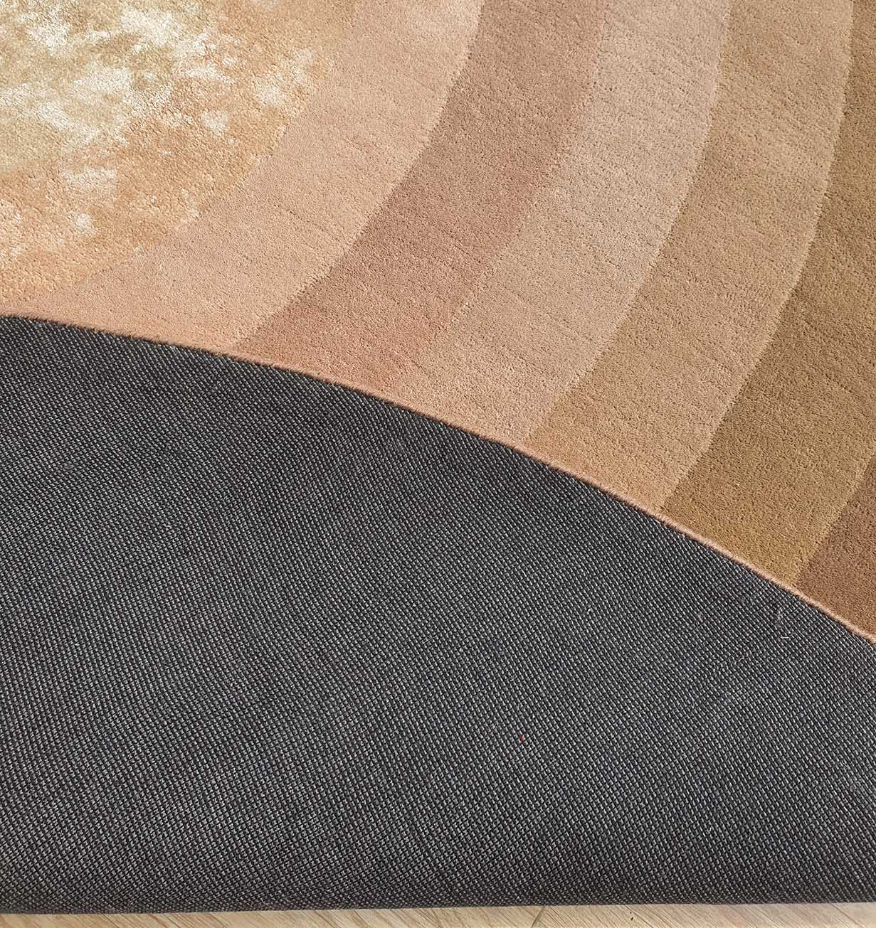 A corner of the Luxurious Wool & Cotton Handmade Rug - HTWV 55 (300cm) by RAMSHA HOME is lifted, showing its textured dark gray underside. The surface displays beige, brown, and peach arcs on a light wood floor.