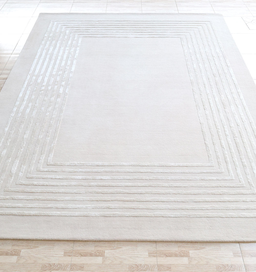 The RAMSHA HOME Hand-Tufted Carpet For Home And Office HTWV-70, a large rectangular beige rug with a raised geometric border of concentric rectangles, is placed on a light wood and tile floor.