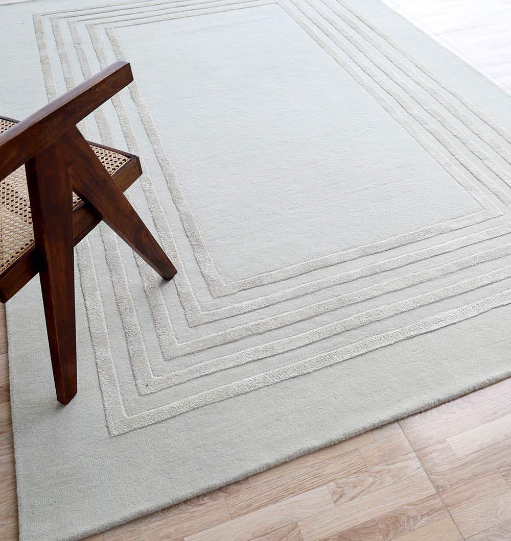 The RAMSHA HOME Hand-Tufted Carpet For Home And Office HTWV-70 features a subtle geometric border and lies on a wooden floor, with the corner of a wooden chair with a woven seat visible at the edge of this modern rug.