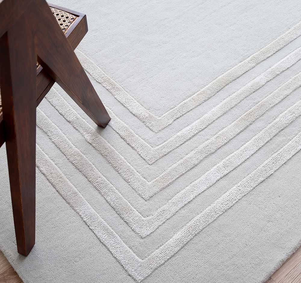 A dark wooden chair with a woven seat rests on the RAMSHA HOME Hand-Tufted Carpet For Home And Office HTWV-70—a light, minimalist wool rug with subtle angular cream patterns.