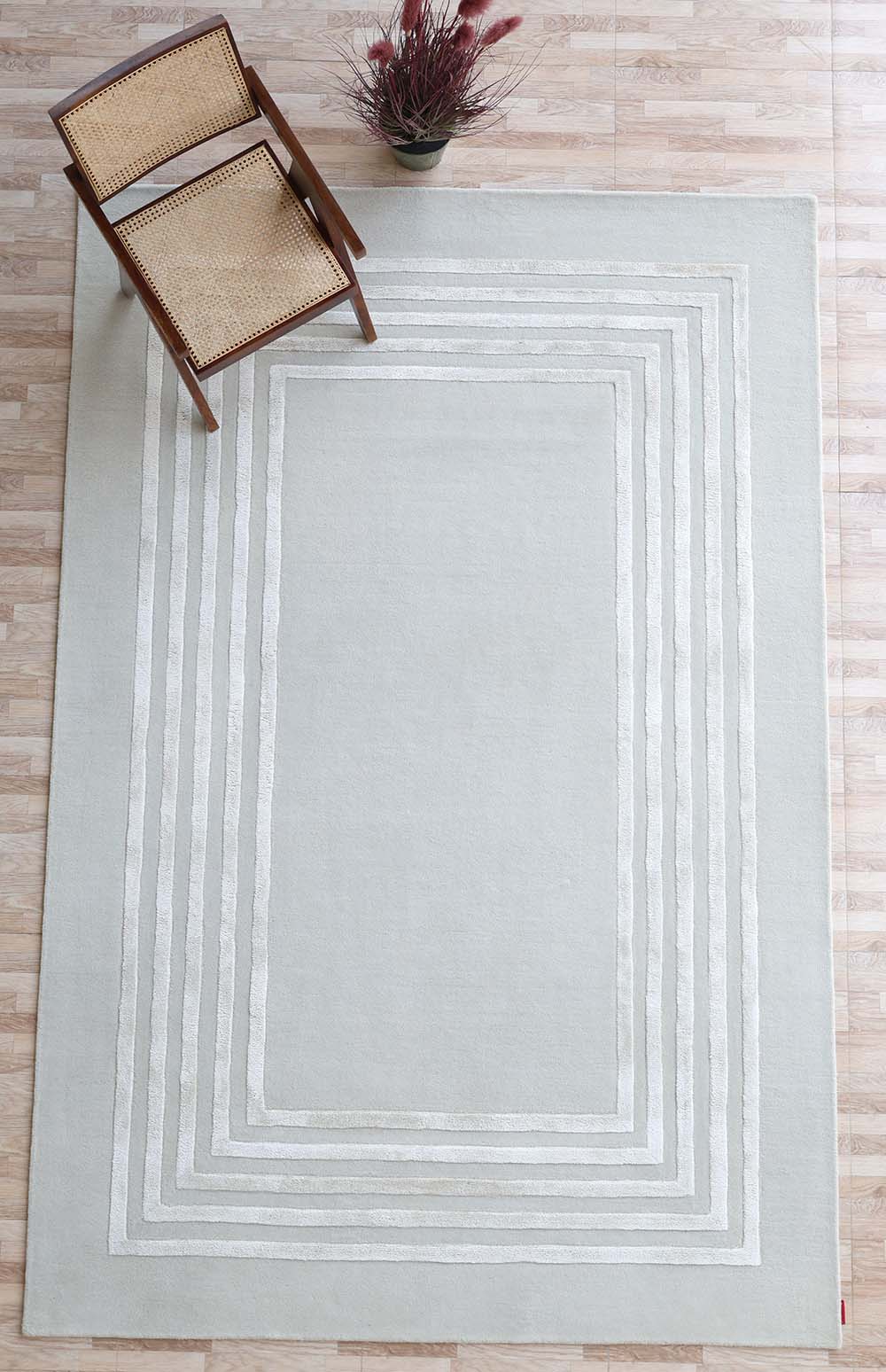 The RAMSHA HOME Hand-Tufted Carpet For Home And Office HTWV-70, in light gray with a geometric border, lays on a light wood floor with a wooden chair featuring a woven cane seat and a potted plant with red-tinted leaves at the top.