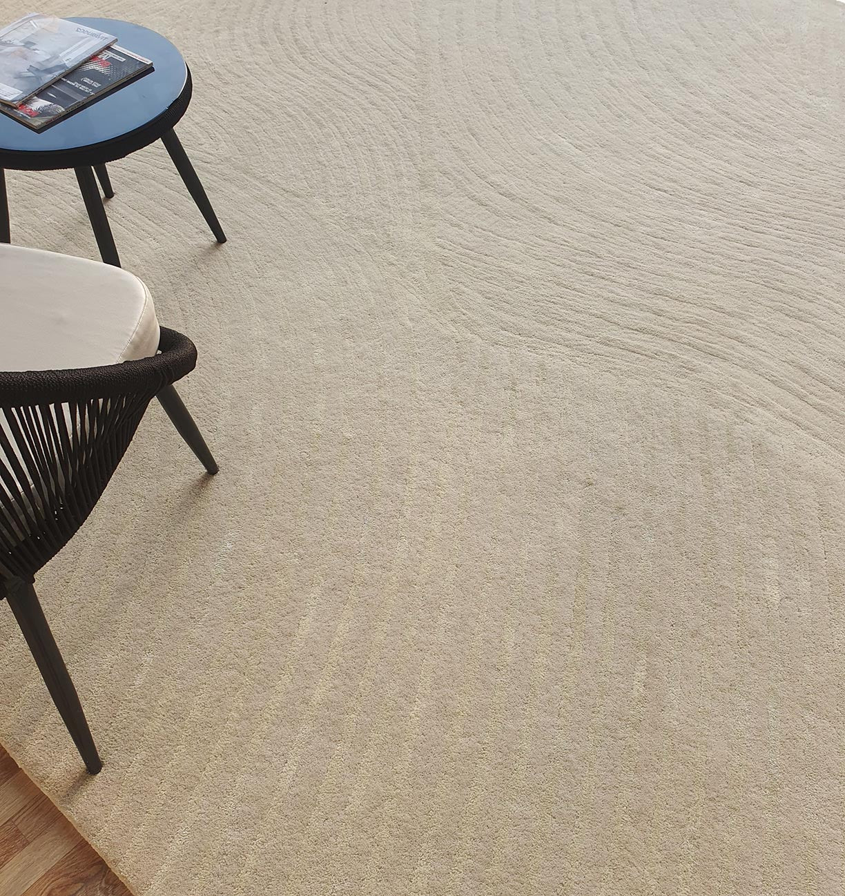 The RAMSHA HOME Luxurious Wool & Cotton Rug - HTWV 71 (300x400cm), featuring textured beige and subtle wavy patterns, lies beneath a black round table with magazines and a chair with a woven black frame and light cushion.