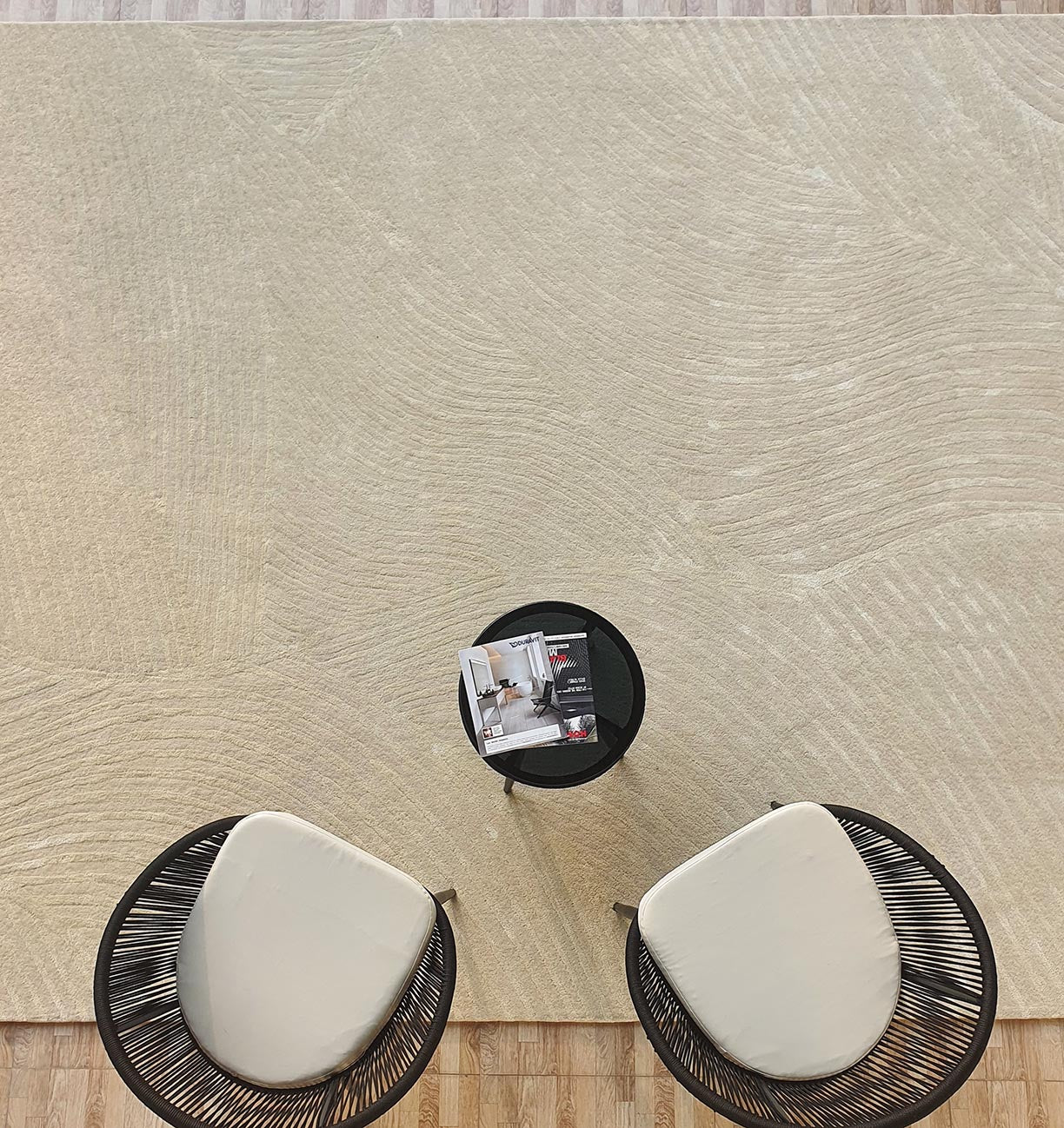 Two modern black chairs with white cushions face a small black round table holding magazines, all set on a RAMSHA HOME Luxurious Wool & Cotton Rug - HTWV 71 (300x400cm), with a wooden floor in the background.