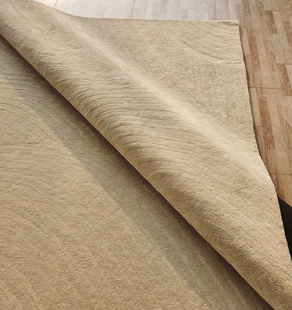 The Luxurious Wool & Cotton Rug - HTWV 71 (300x400cm) by RAMSHA HOME is shown partially folded, highlighting its texture and underside as it rests on a wooden floor with contrasting light and dark grain.