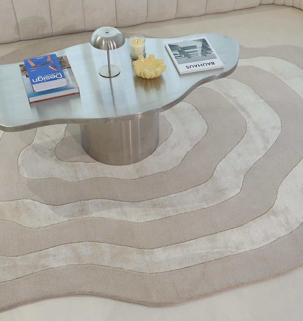 A wavy-edged coffee table with a metallic base sits on the RAMSHA HOME Wool & Tencel Multi Color Rug HTWV-91. On the table are books, a small lamp, a yellow candle, and a decorative shell-shaped object.