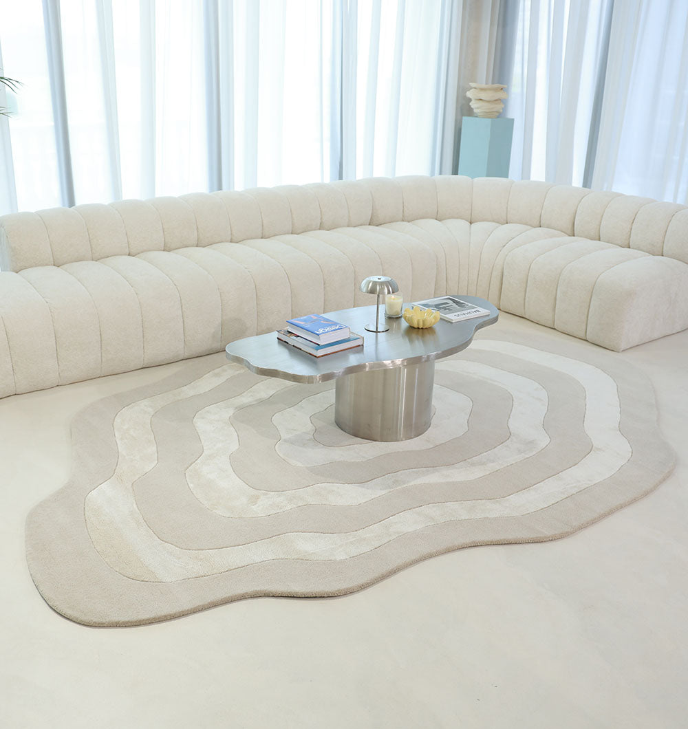 A modern living room with a cream curved sectional, the RAMSHA HOME Wool & Tencel Multi Color Rug HTWV-91, a metallic sculptural coffee table with books and decor, and sunlight filtering through sheer curtains.