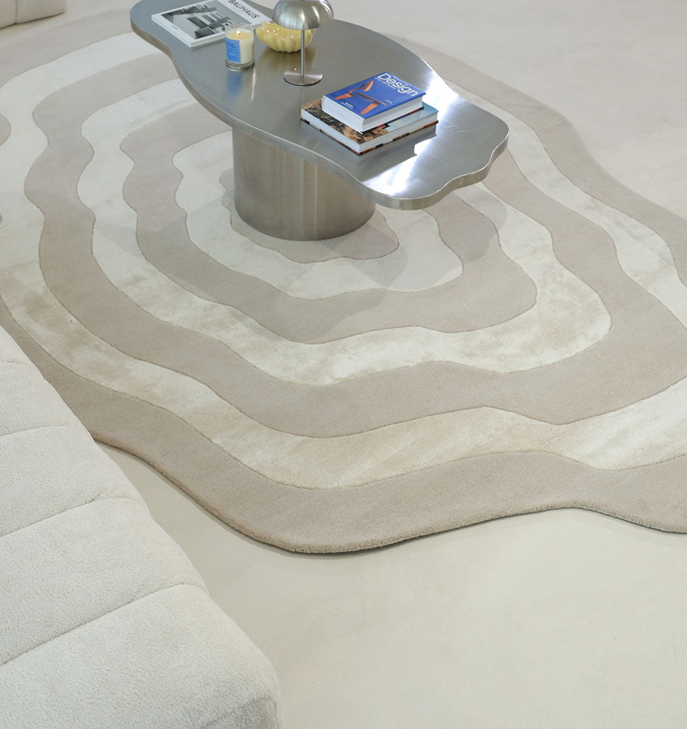 A modern living room features the RAMSHA HOME Wool & Tencel Multi Color Rug HTWV-91 in wavy beige and cream, a metallic coffee table with books and decor, and a cream sofa atop a light floor.