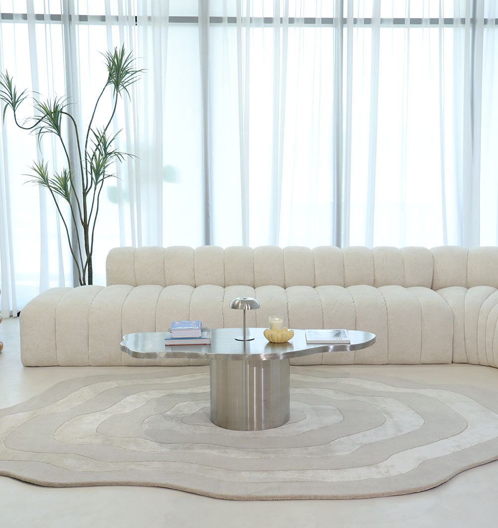 A modern living room features a wavy cream sofa, metallic coffee table with books and décor, a lamp, and is anchored by the RAMSHA HOME Wool & Tencel Multi Color Rug HTWV-91, with sheer curtains and a tall plant in the background.
