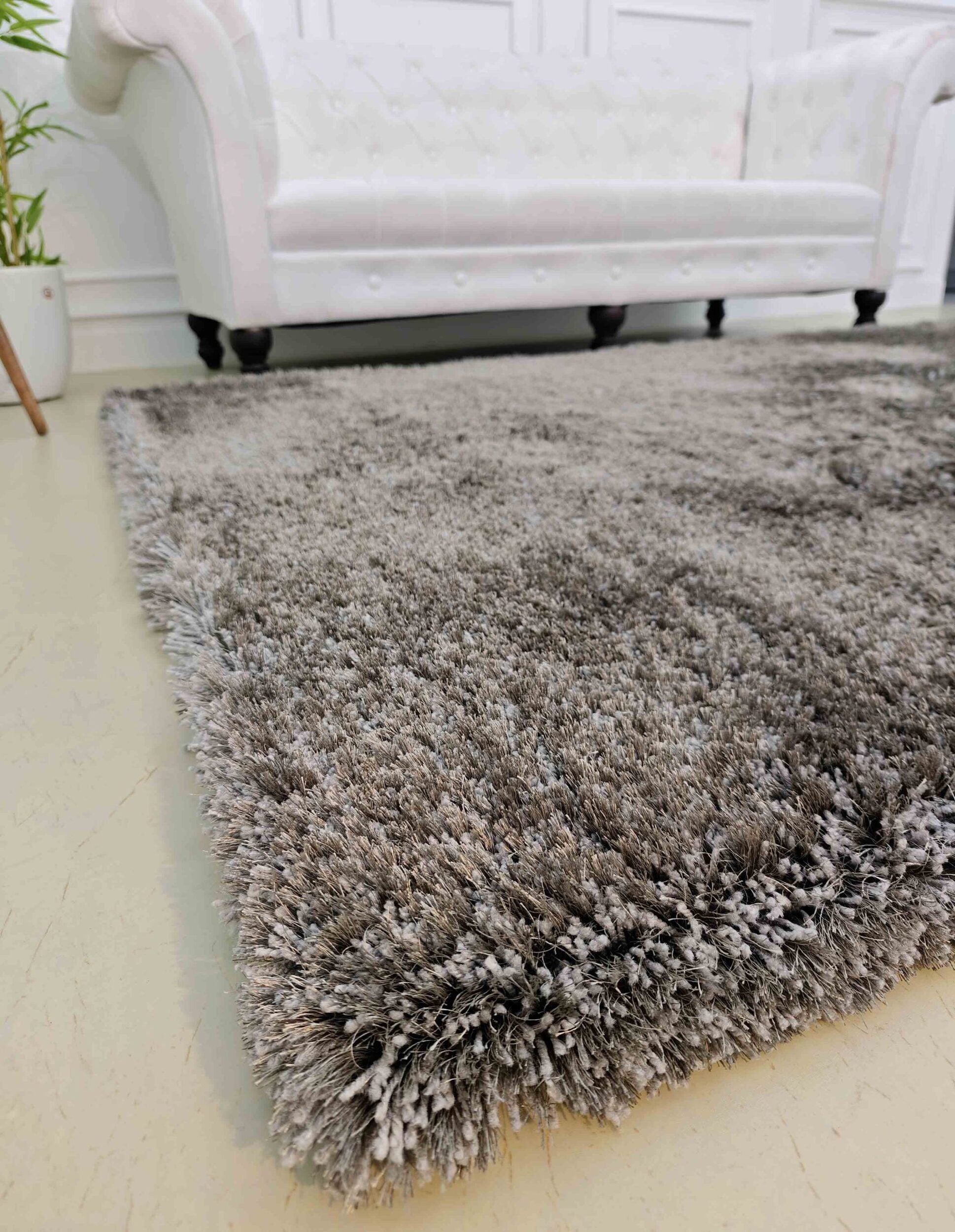 A close-up of the RAMSHA HOME Grey Color Rectangle Shaped Shaggy Rug - SG 05 on a light floor, with a white tufted sofa and potted plant in the background.