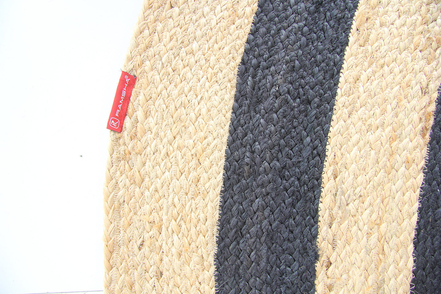 A close-up of the RAMSHA HOME Handmade Braided Jute Floor Round Rug - BR 058 (200cm) shows alternating beige and dark brown stripes, with a small red fabric tag displaying white text attached to the beige section on the left.
