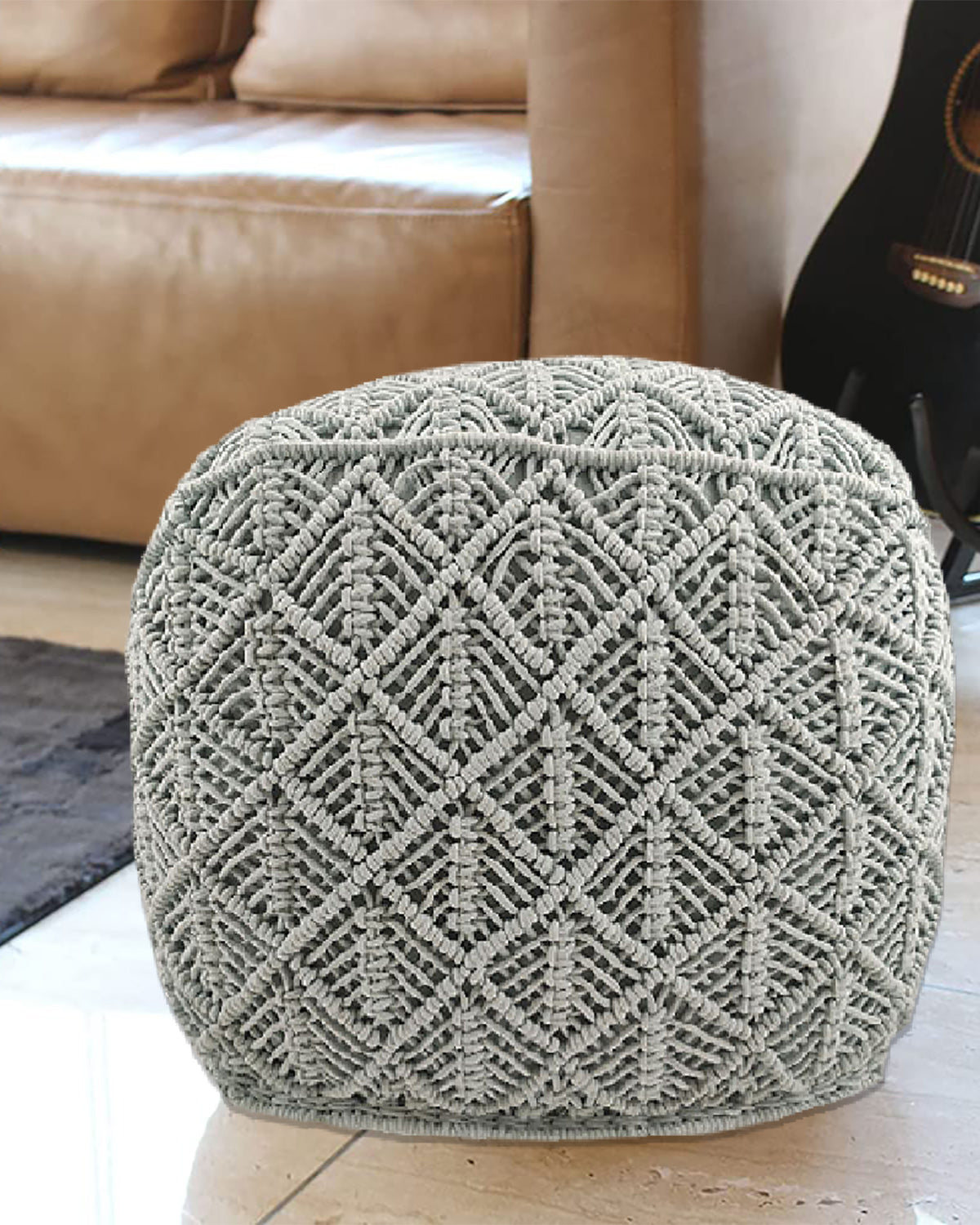 The RAMSHA HOME Macrame Cotton Pouf, featuring textured light gray cotton and a diamond pattern, sits on a tile floor beside a tan leather sofa and black guitar, infusing the living room with boho-style charm.