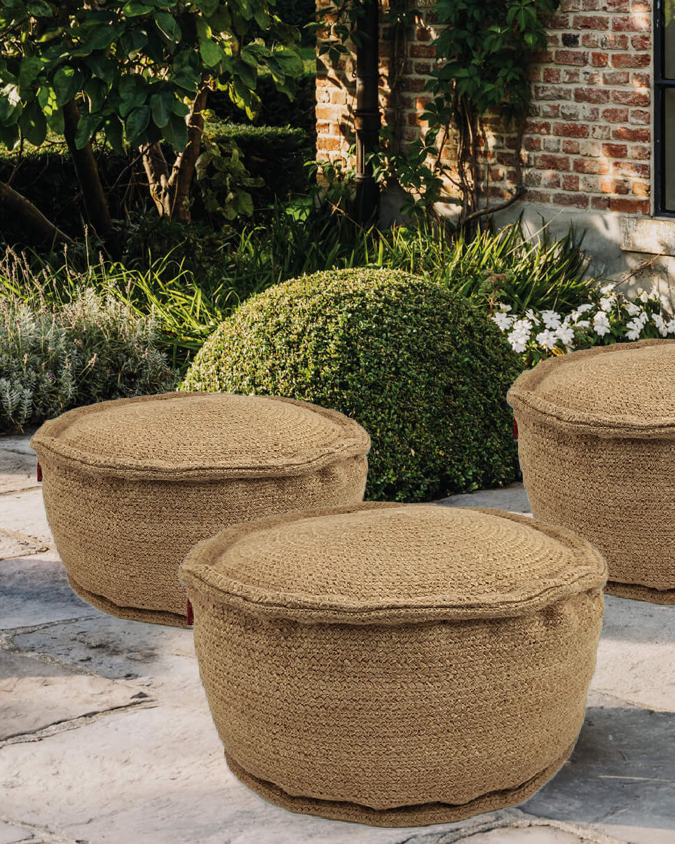 Three Natural Color Jute Pouf PF-056 by RAMSHA HOME sit on a stone patio in a garden, providing eco-friendly outdoor seating with green bushes, plants, and a brick building in the background.