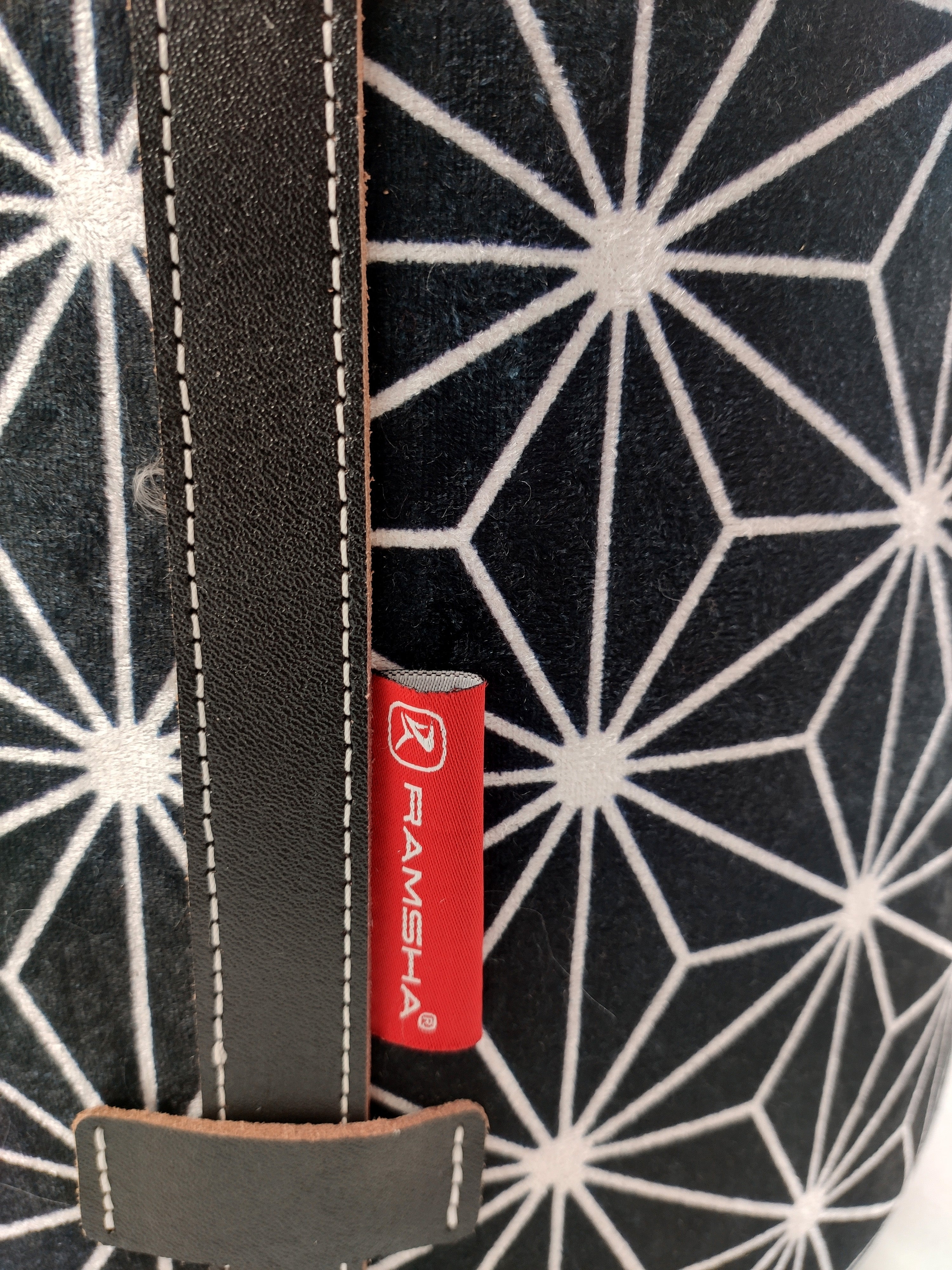 Close-up of the Printed Pedestal Pouf With Leather Handle PF-027 by RAMSHA HOME, featuring a black fabric with white geometric stars, a vertical leather handle, and a red RAMSHA HOME tag—ideal for stylish home decor accents.
