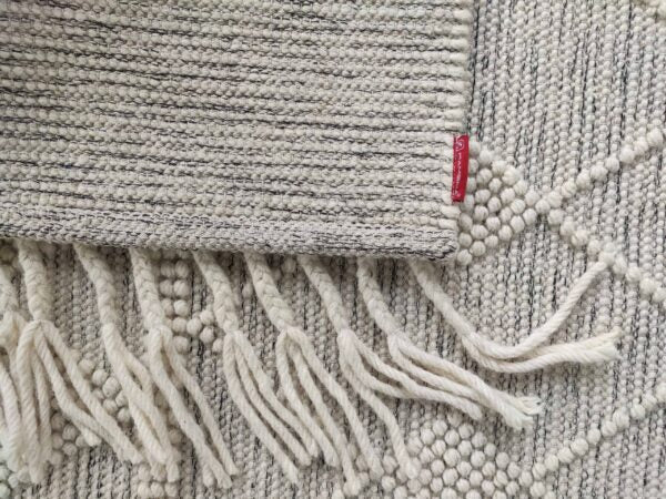 A close-up of the RAMSHA HOME Fringes Woven Rug For Indoor, Floor Rug SRN-017 shows its textured wool design with fringe edges, partially atop a cream and grey diamond-patterned rug. A small red fabric tag is attached to the fringed rug.