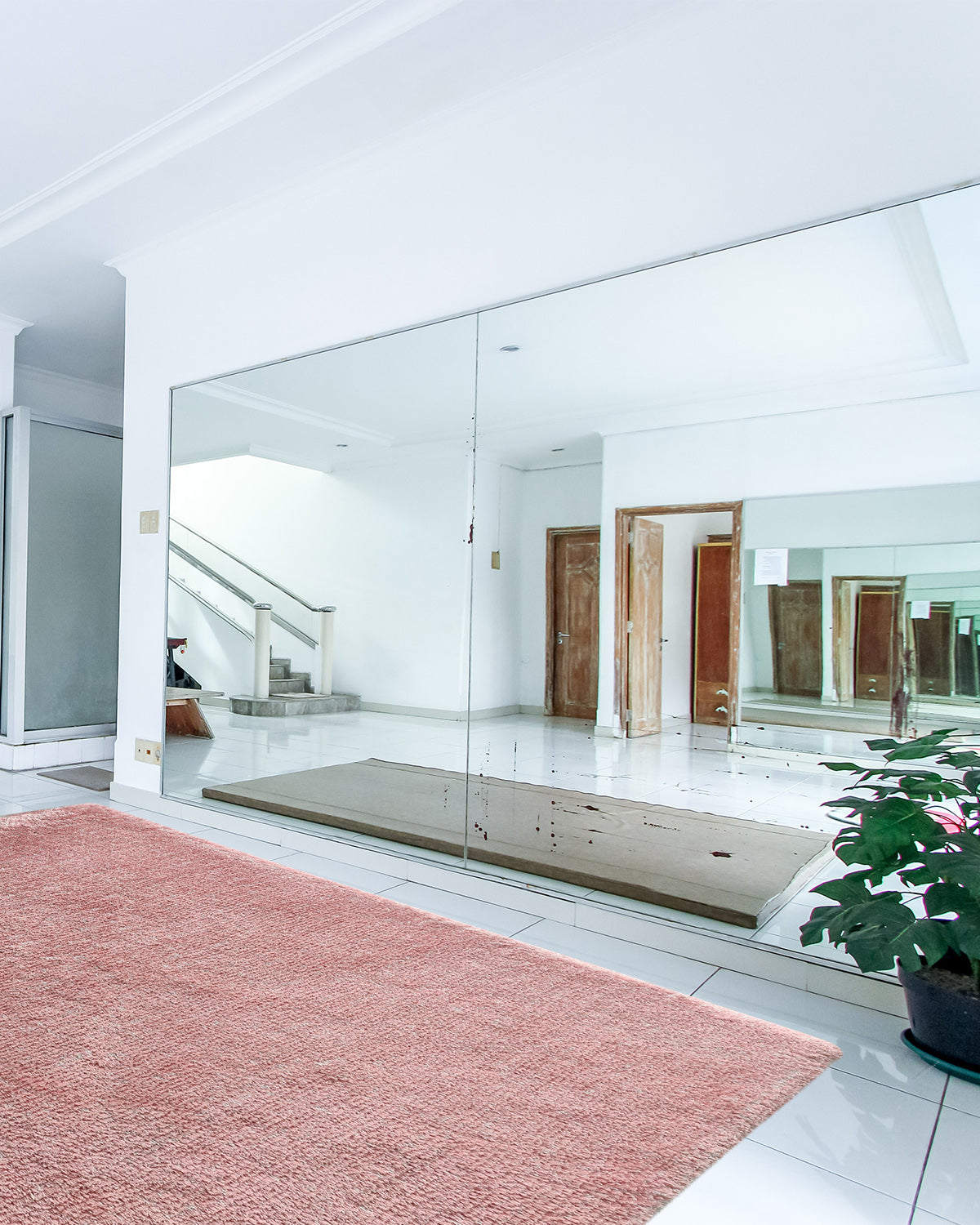 A spacious room with a white tiled floor features the RAMSHA HOME Pink Color Modern Kids Room Decor Shaggy Rug (MPV 06, 200x300cm), a potted plant, and mirrored walls reflecting doors, stairs, and a bright interior.