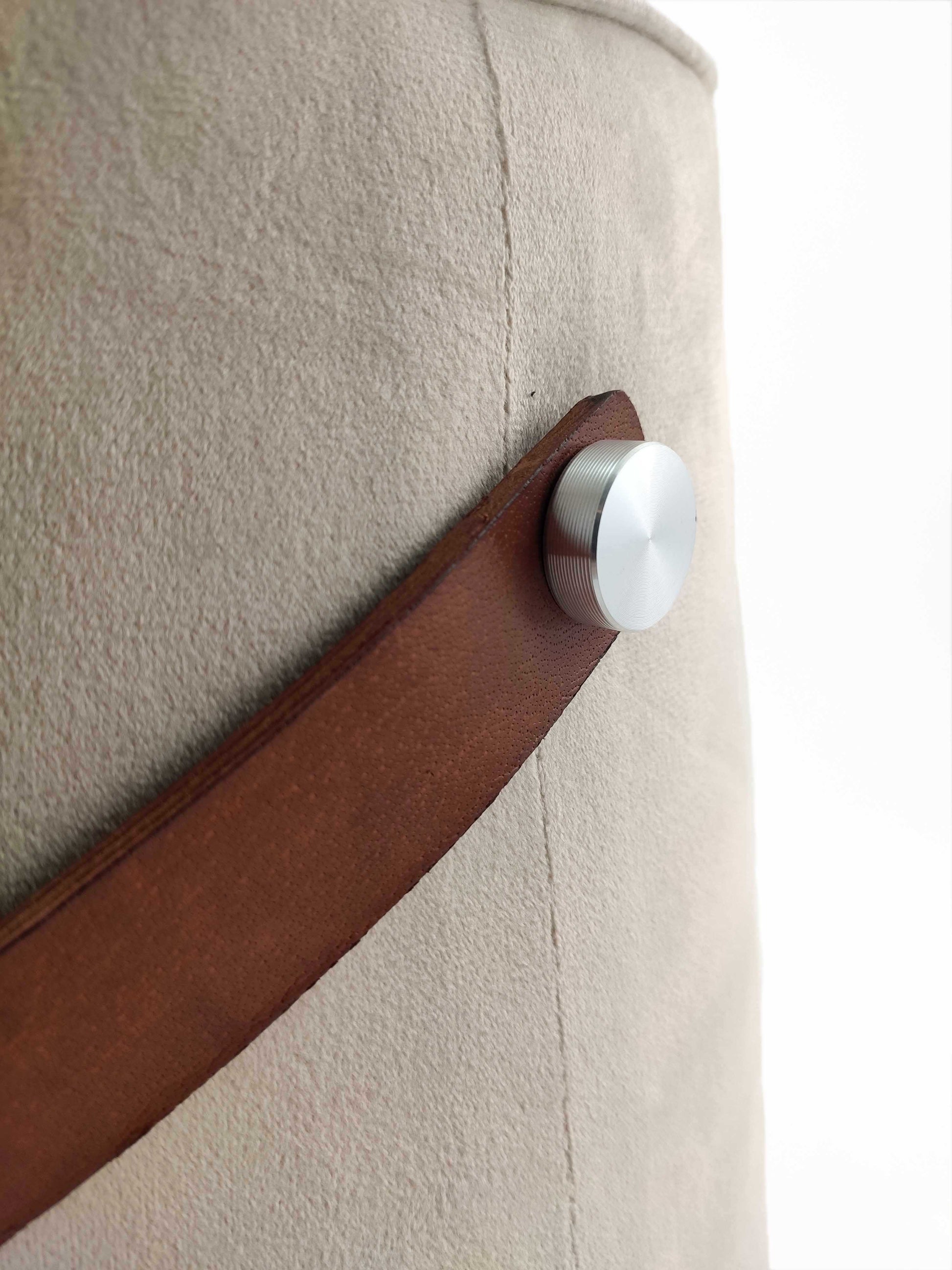 Close-up of a beige suede surface with a brown leather handle attached by a round silver knob highlights the minimalist, modern design of the Leather Pedestal Pouf With Leather Handle by RAMSHA HOME.