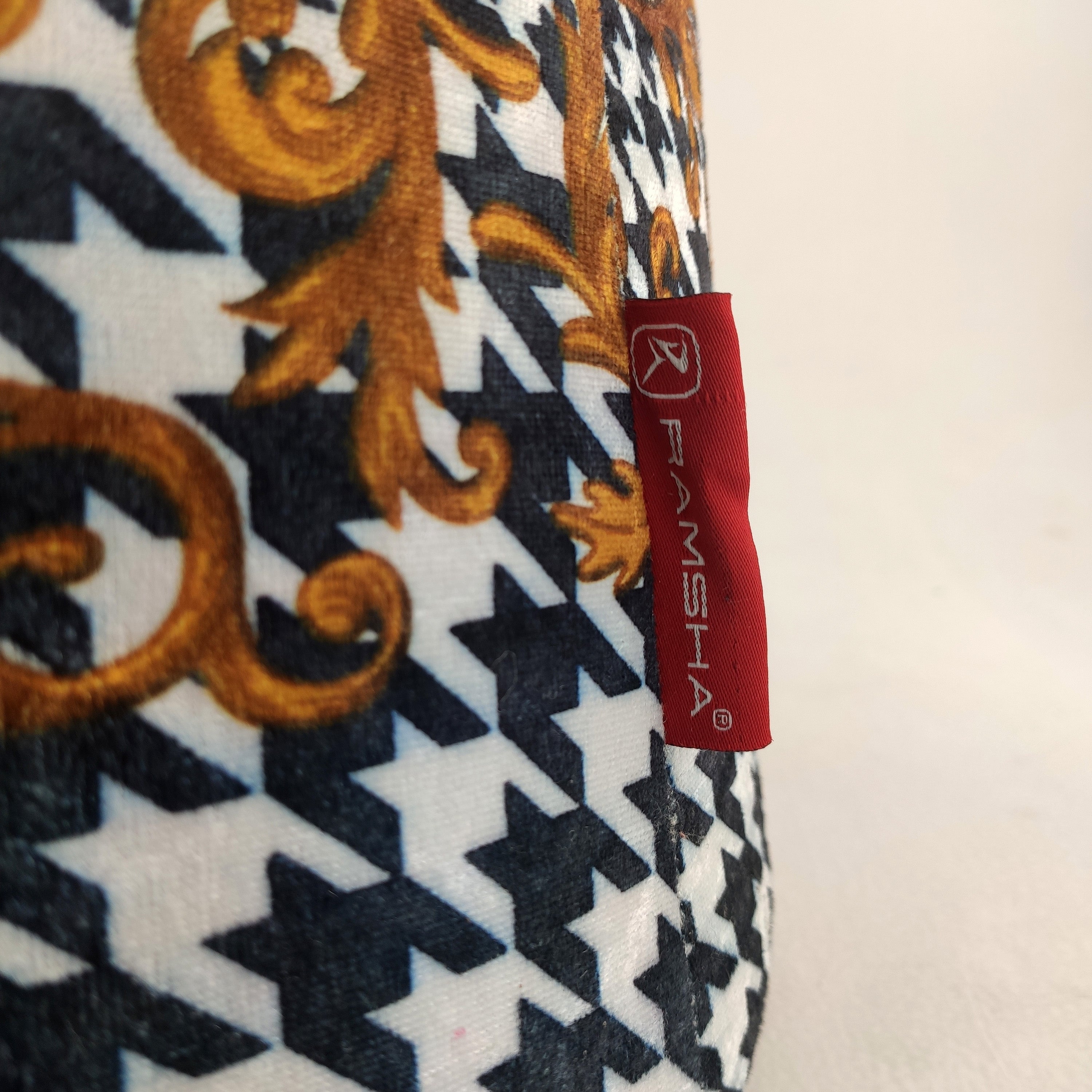 A close-up of a patterned fabric on the RAMSHA HOME Leather Printed Pedestal Pouf With Leather Handle PF-025, featuring black and white houndstooth, gold baroque designs, and a red RAMSHA® tag—ideal as a chic footrest or accent pouf.
